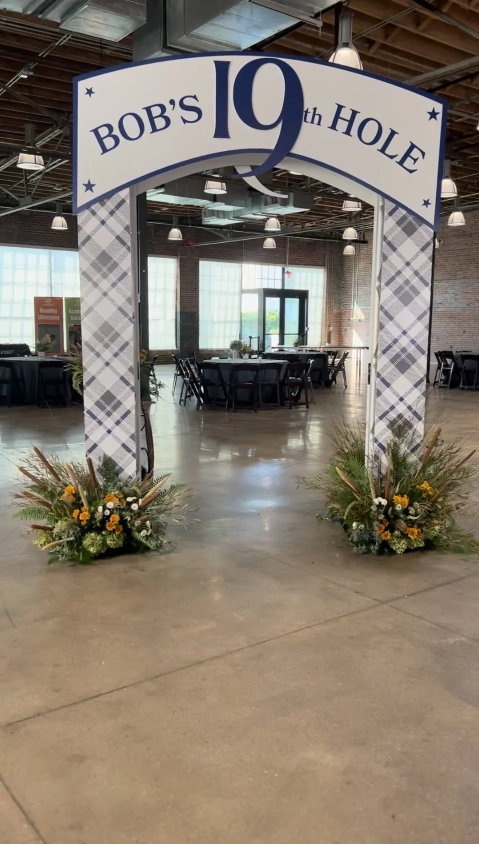 Decorative arch with a sign that reads "Bob's 19th Hole" and floral arrangements at the base, set up indoors in a large, industrial-style venue with tables and chairs in the background.