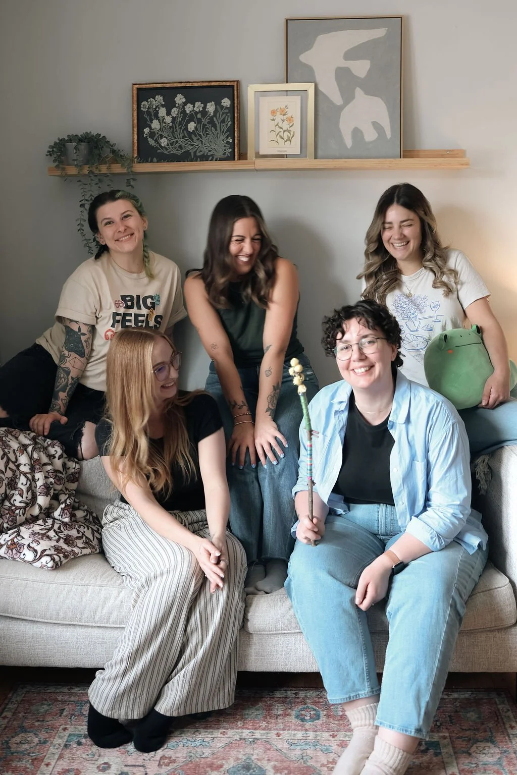 Group of six women in a living room, smiling and laughing, sitting on and around a beige sofa, with artwork and a plant on the wall behind them.