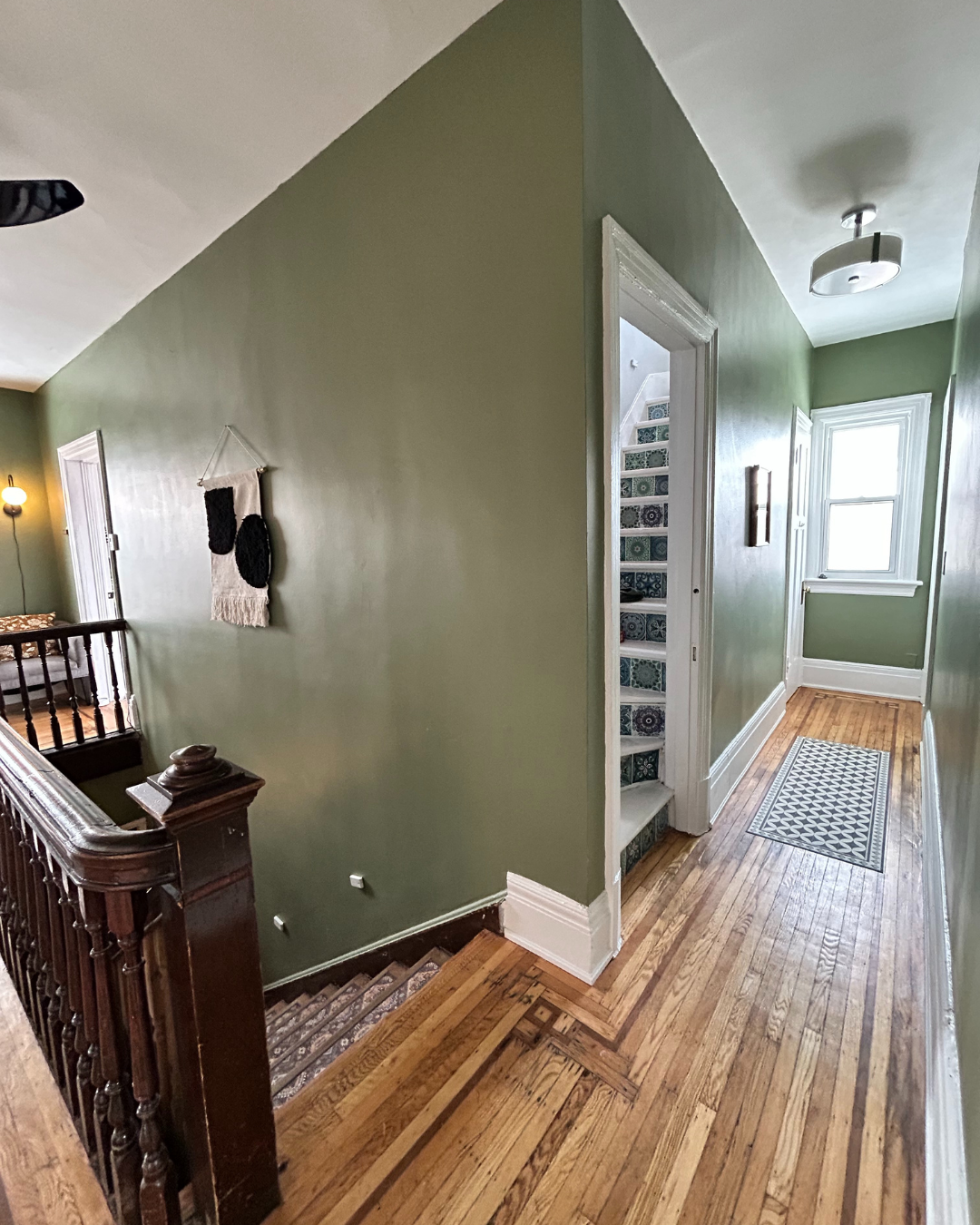Interior view of a hallway with wooden floors, green walls, a staircase with a carpet runner, and a window at the end of the hallway.