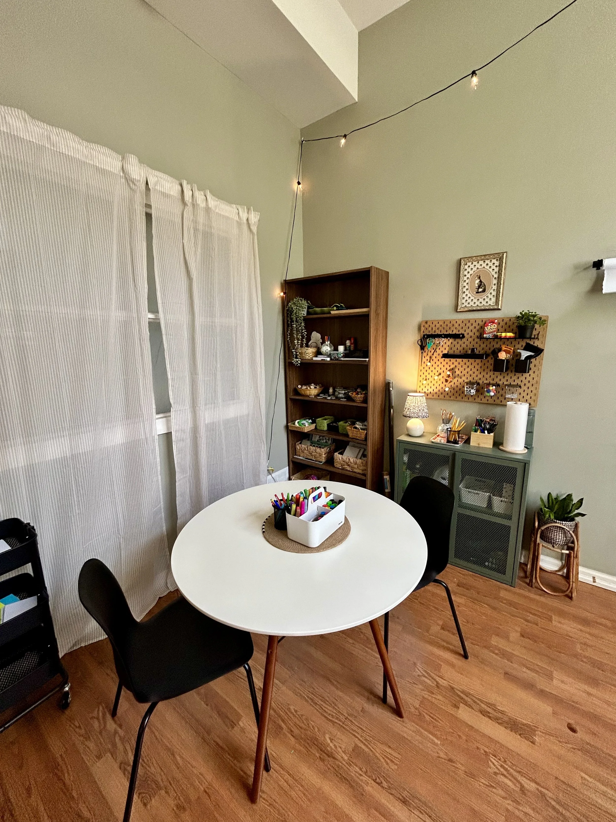 A cozy craft room with a white round table, black chairs, a wooden bookshelf, a green cabinet, and a pegboard with supplies. String lights are hanging from the ceiling, and there are plants and art on the walls.