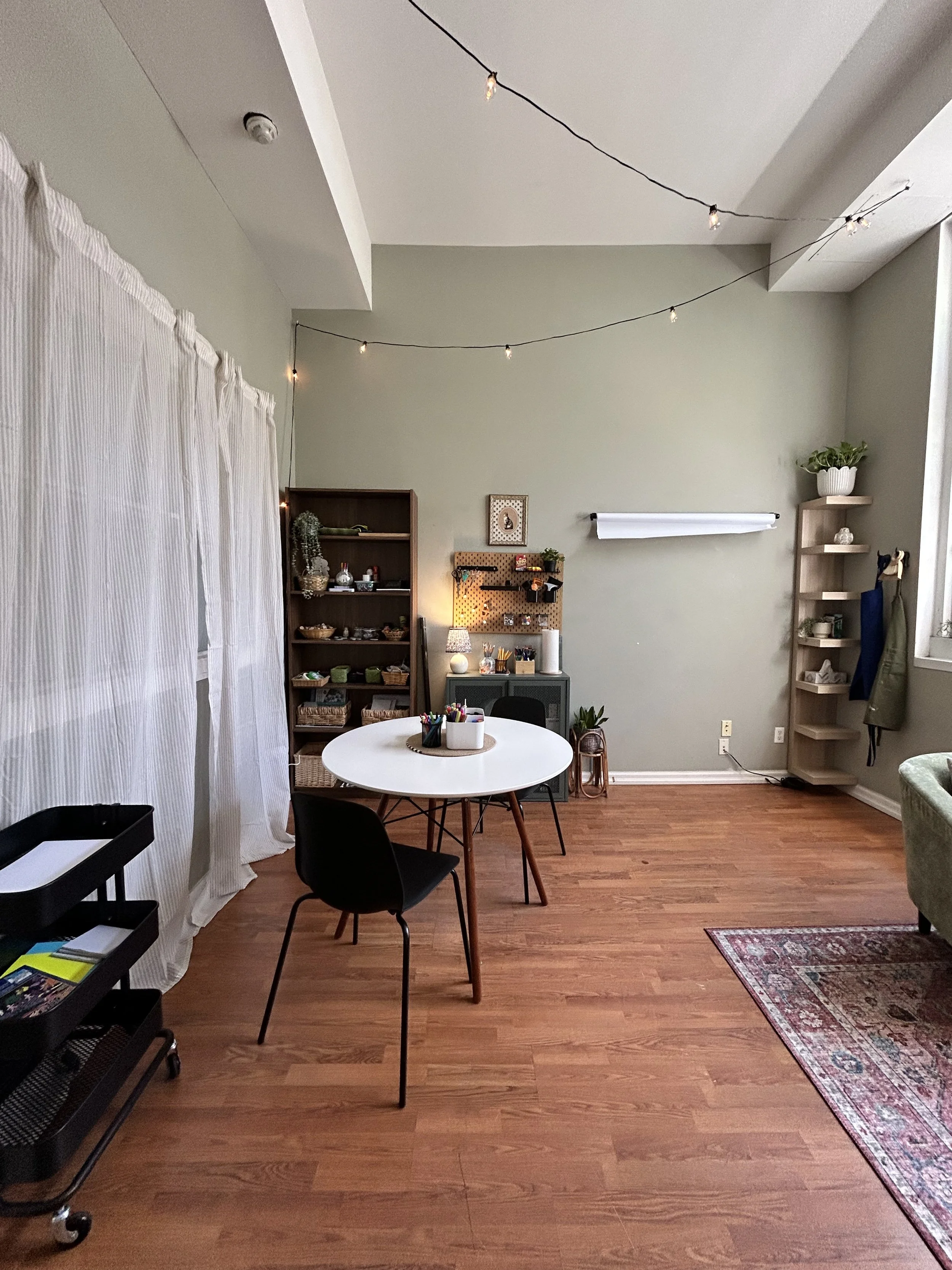 A room with hardwood floors, a white round table with black chairs, a shelving unit, fairy lights on the ceiling, and a window with sheer curtains.