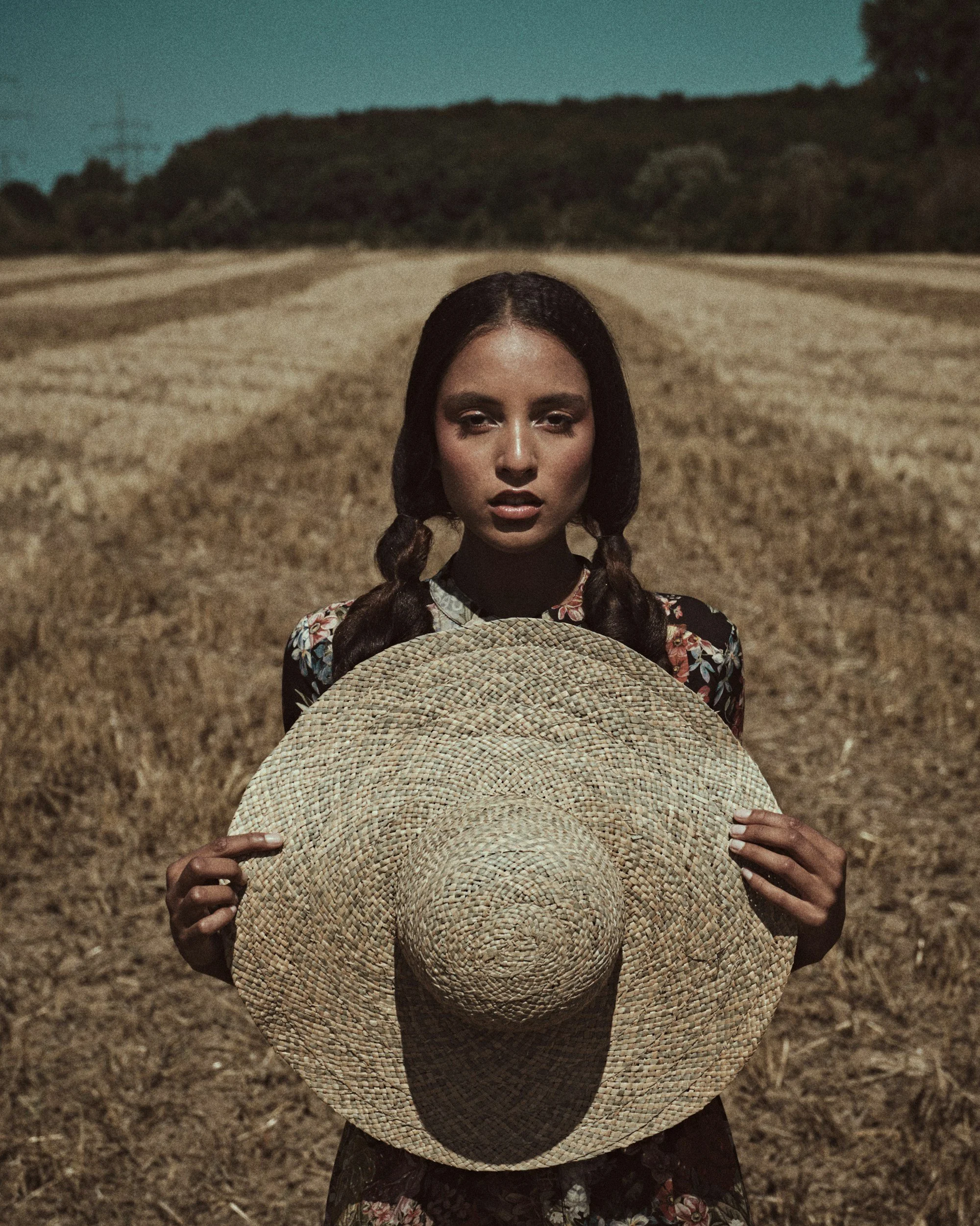 Young woman with braided hair holding a large straw hat, standing in a harvested field with hills and trees in the background.