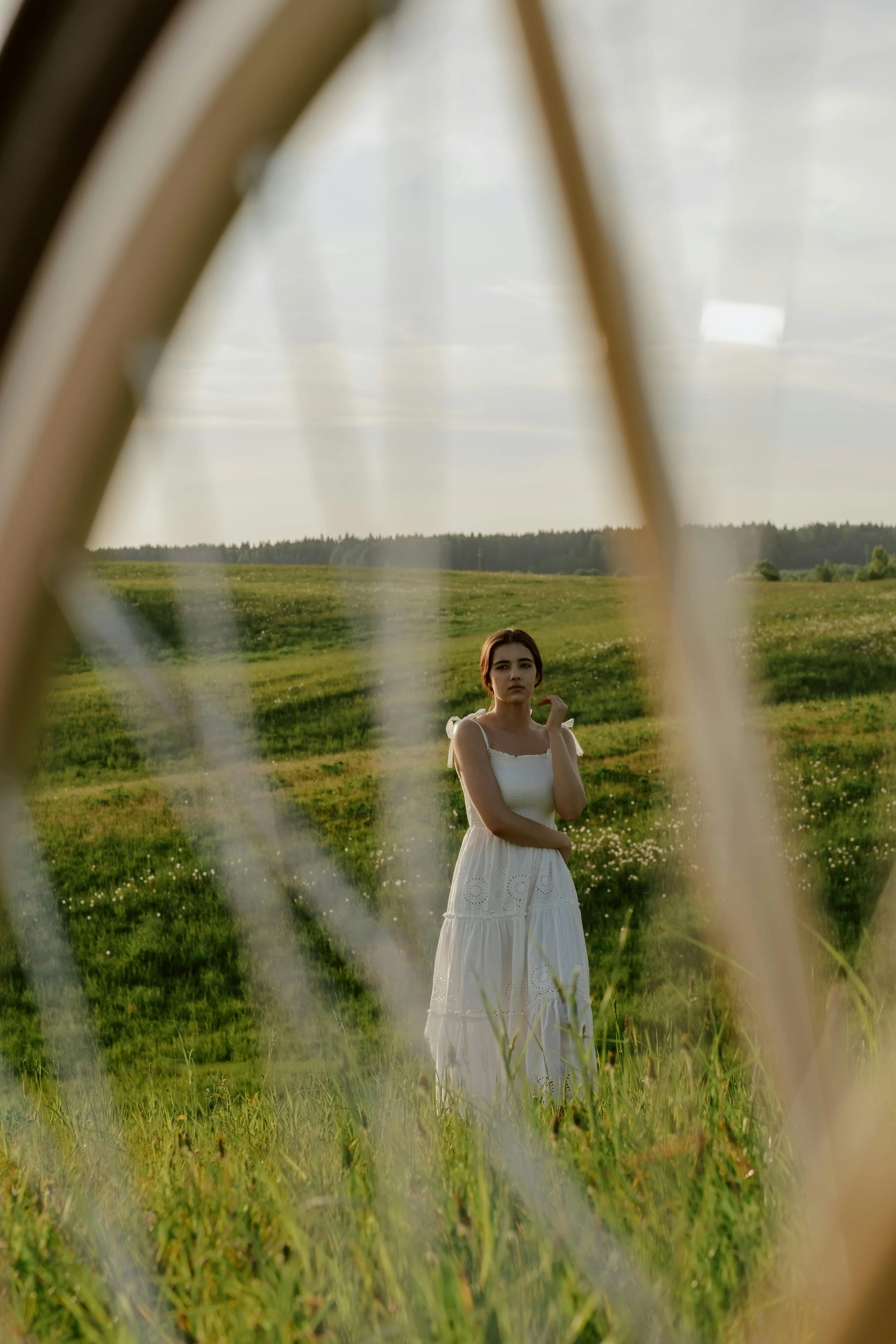 A woman in a white dress standing in a green field, viewed through the spokes of a bicycle wheel.