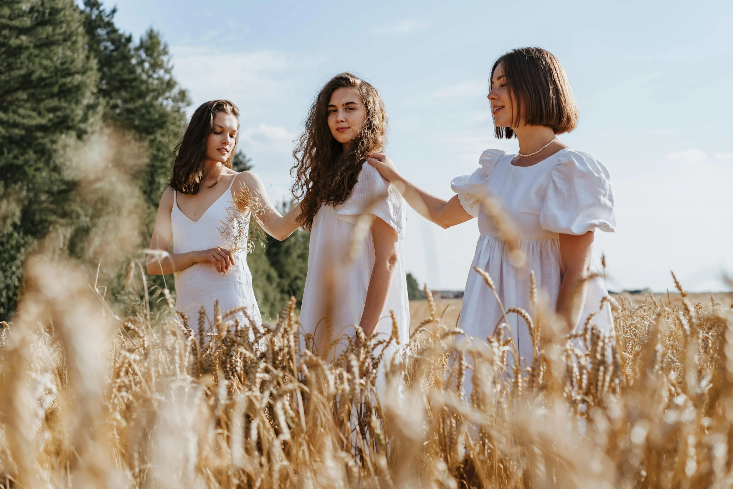 Three young women in white dresses standing in a wheat field during daytime, with one woman touching the shoulder of another.