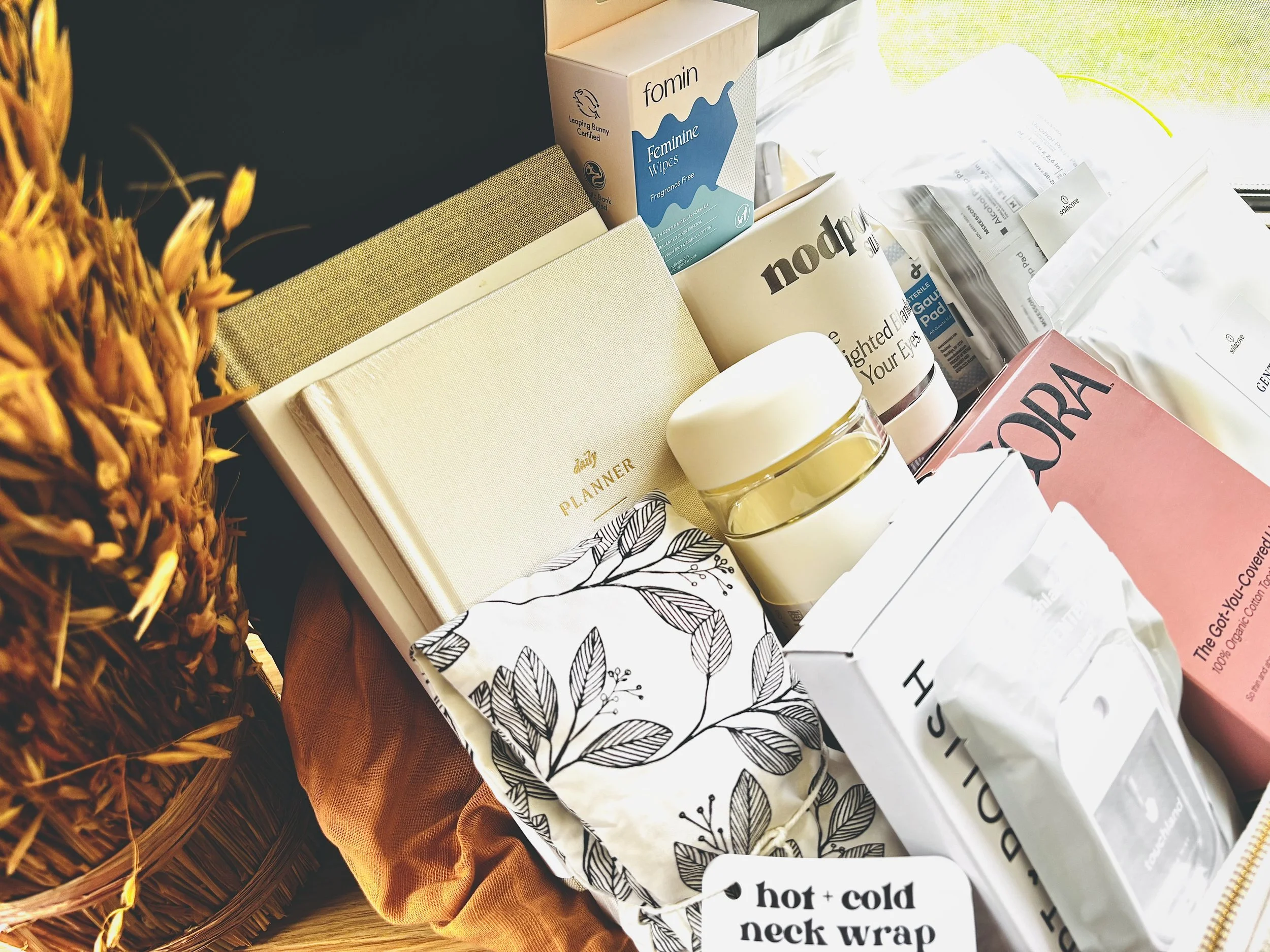A collection of skincare and wellness products, including a box of feminine wipes, a jar of cream, herbal supplements, a planner, candles, and a hot and cold neck wrap, arranged on a surface next to a basket of dried wheat.