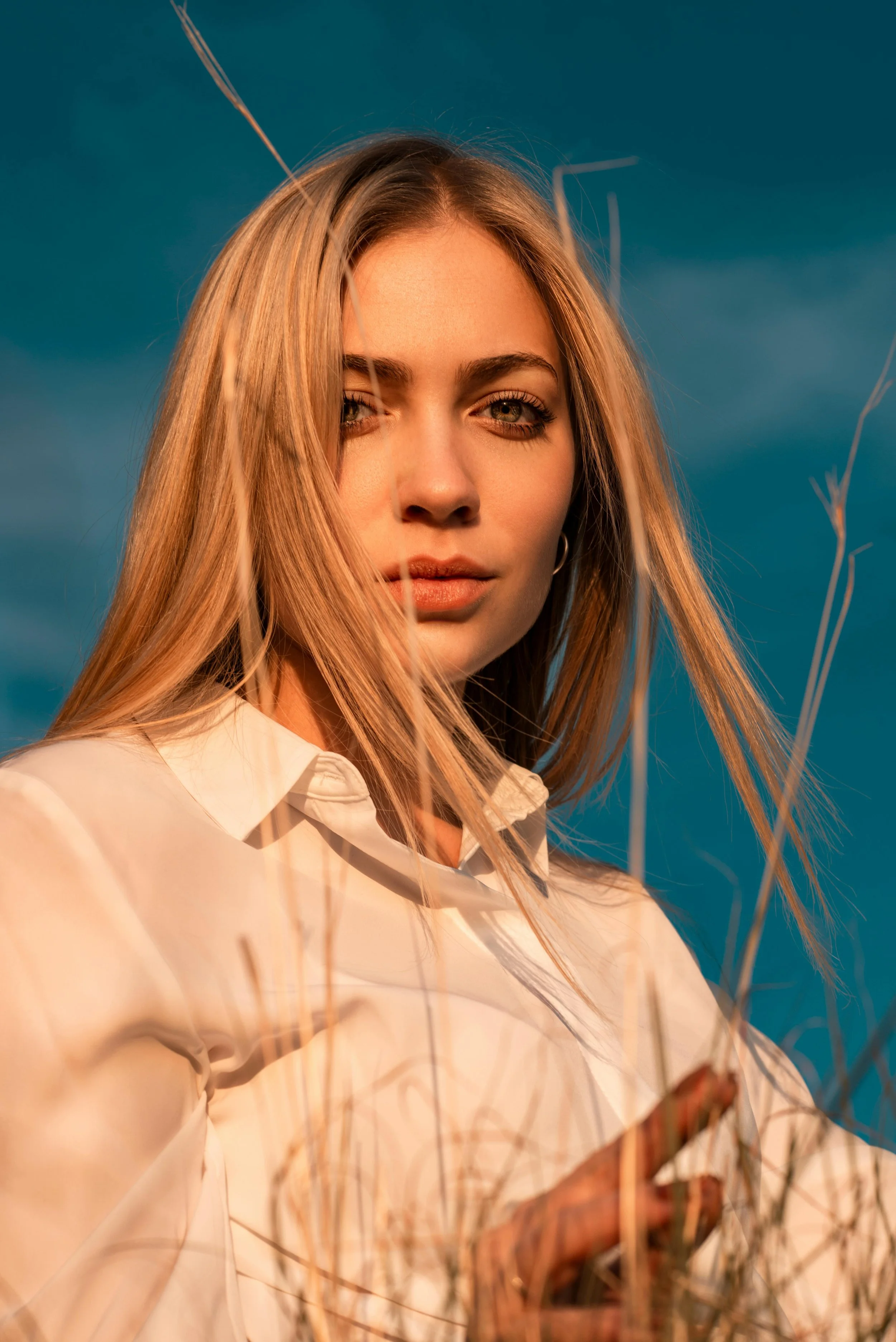 A young woman with blonde hair and blue eyes wearing a white shirt standing outdoors among dry grass, with a cloudy sky in the background, lit by warm sunlight.