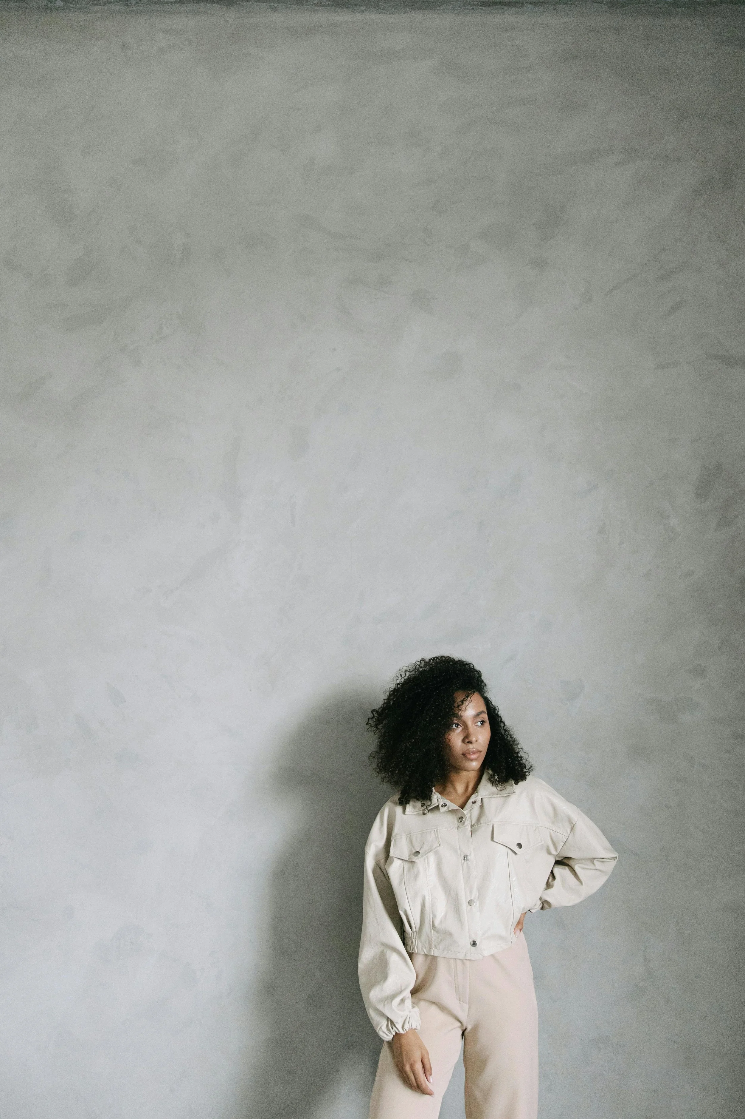 A woman with curly black hair wearing a beige jacket and beige pants standing against a textured light gray wall