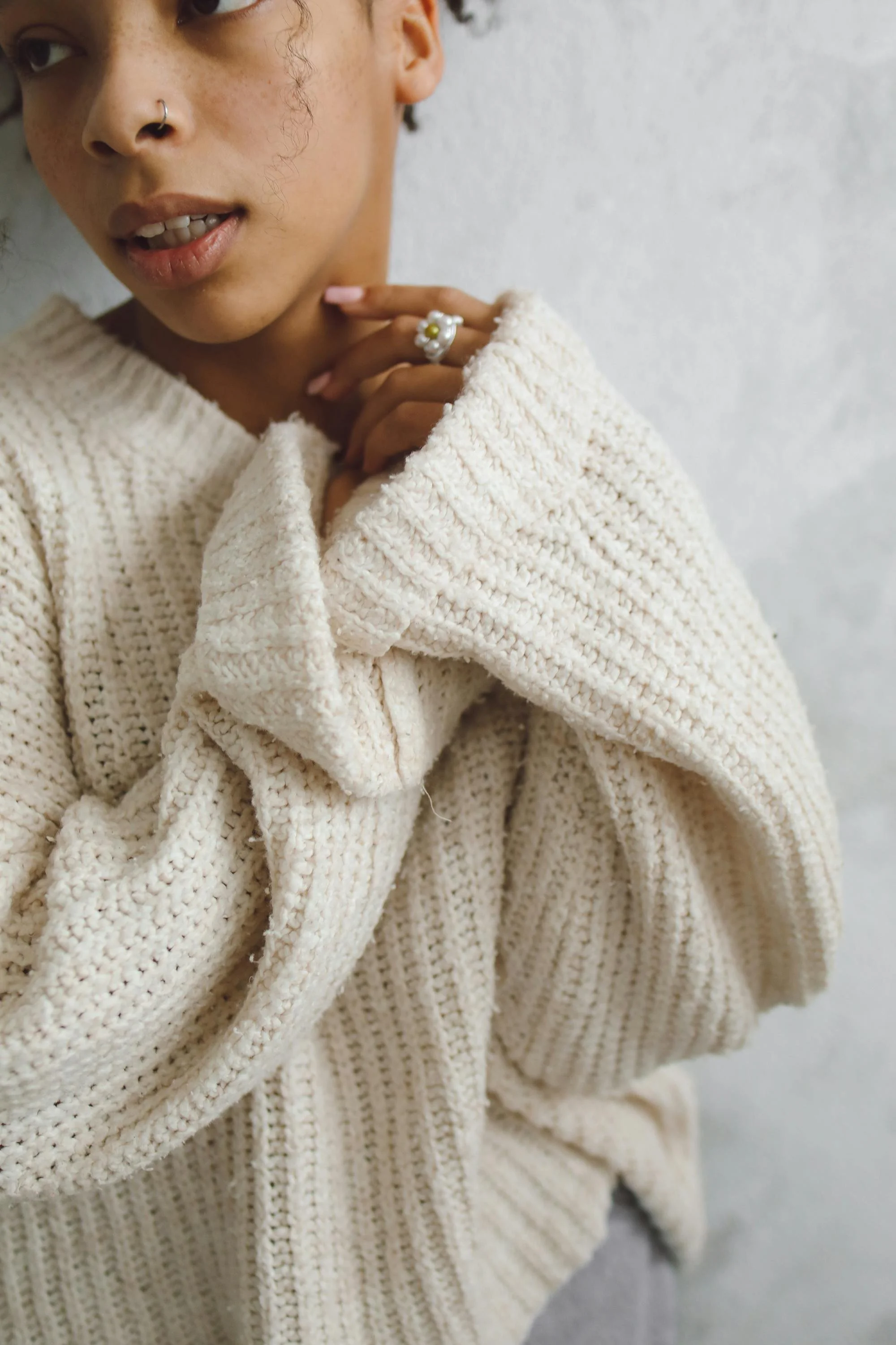Close-up of a woman wearing a chunky cream-colored knit sweater, with her hand touching her neck and a ring with pearls on her finger, against a plain white wall.