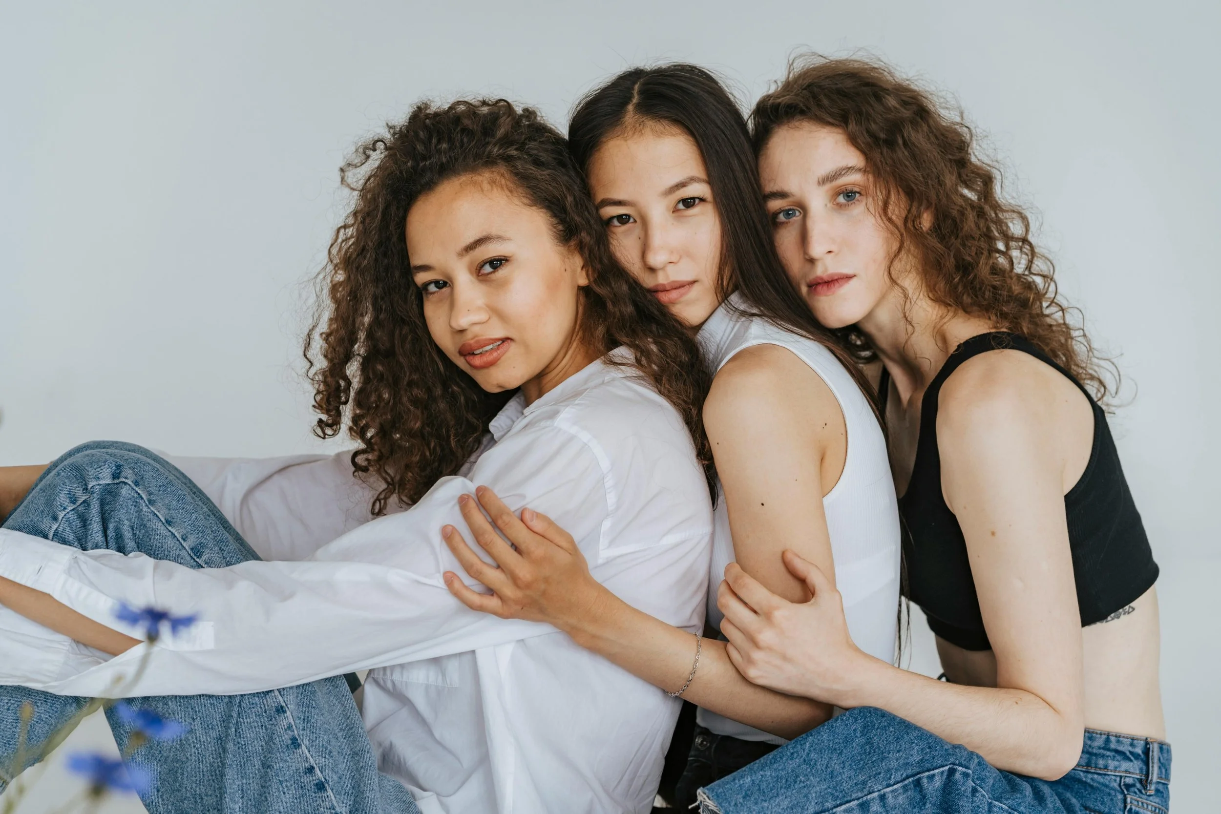 Three young women sitting close together, holding each other and looking at the camera, with light hair and curly hair, against a plain background.