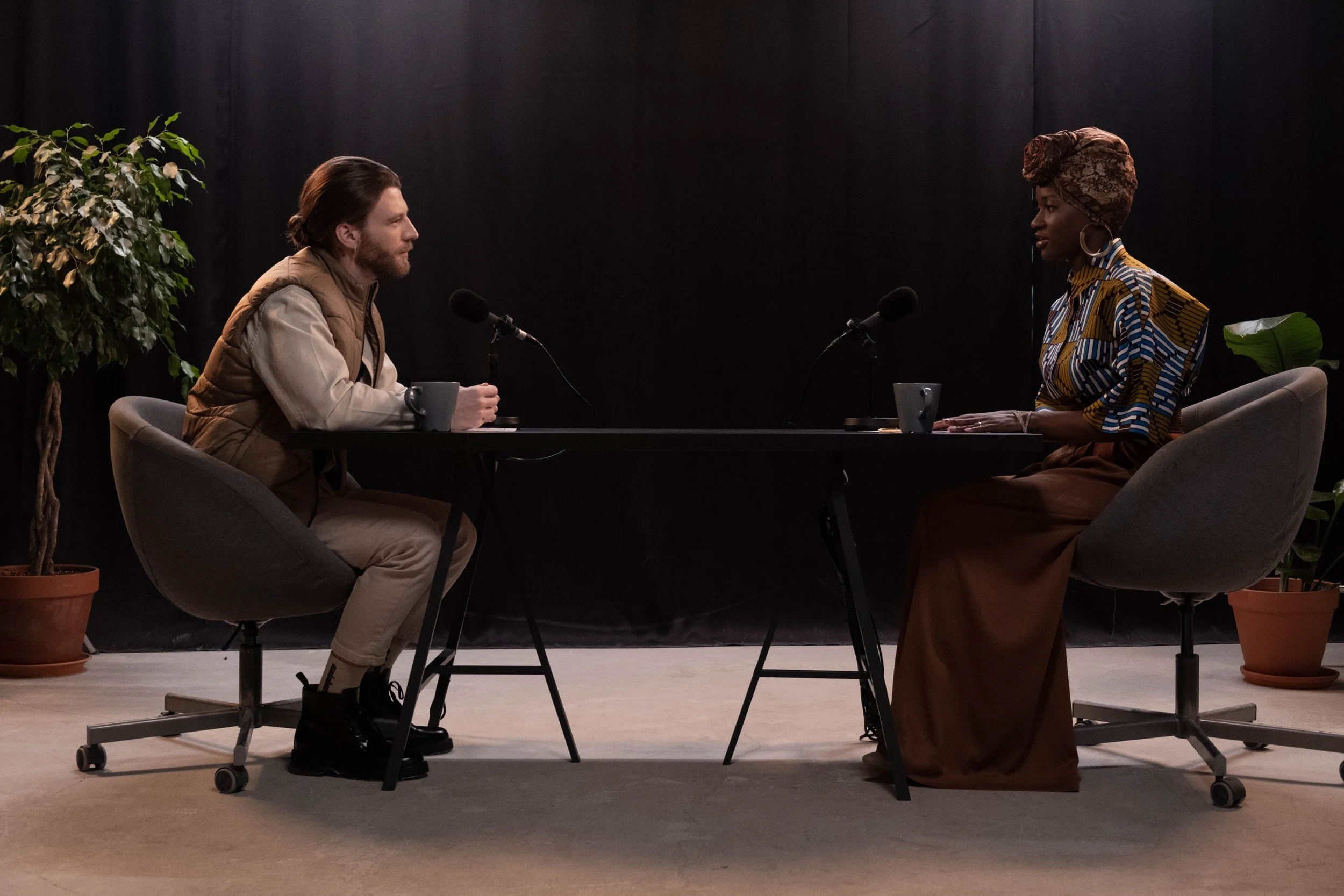 A man and woman sitting across from one another at a table, each with a microphone and coffee mug.
