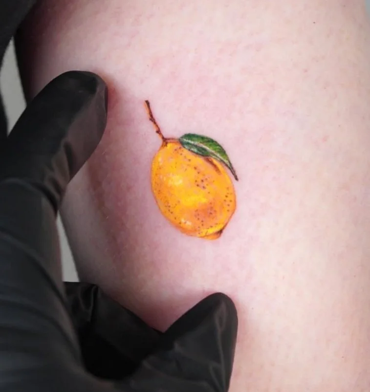 Tattoo of a yellow lemon with a green leaf and brown stem on skin.