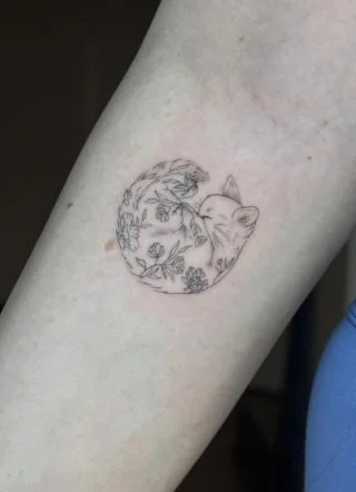 Tattoo of a fox curled in a circle surrounded by floral designs on skin.