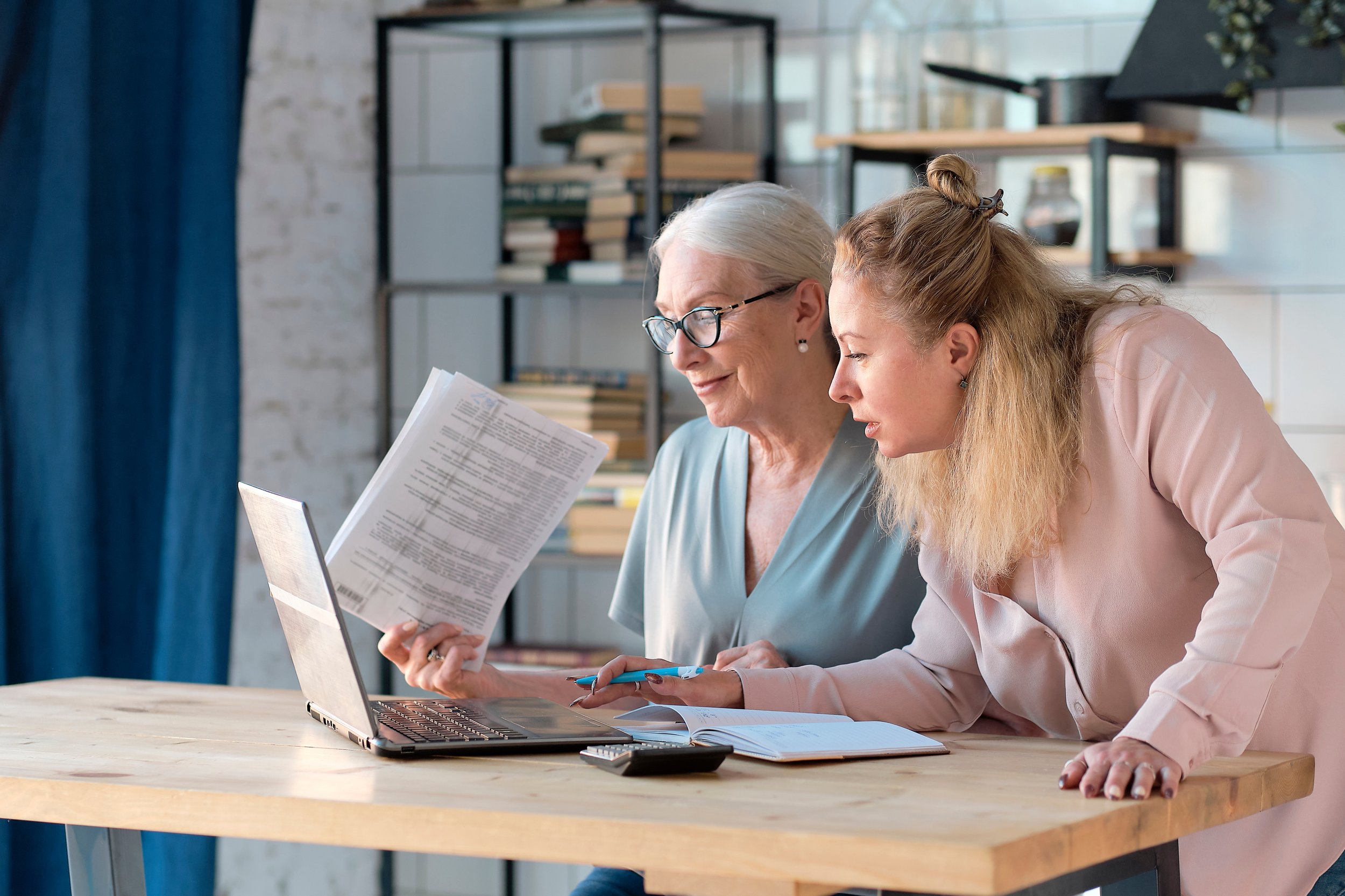 An elderly woman with gray hair and glasses sitting at a table with a younger woman with blonde hair. They are looking at a laptop and documents together in a modern room with bookshelves and window in the background.