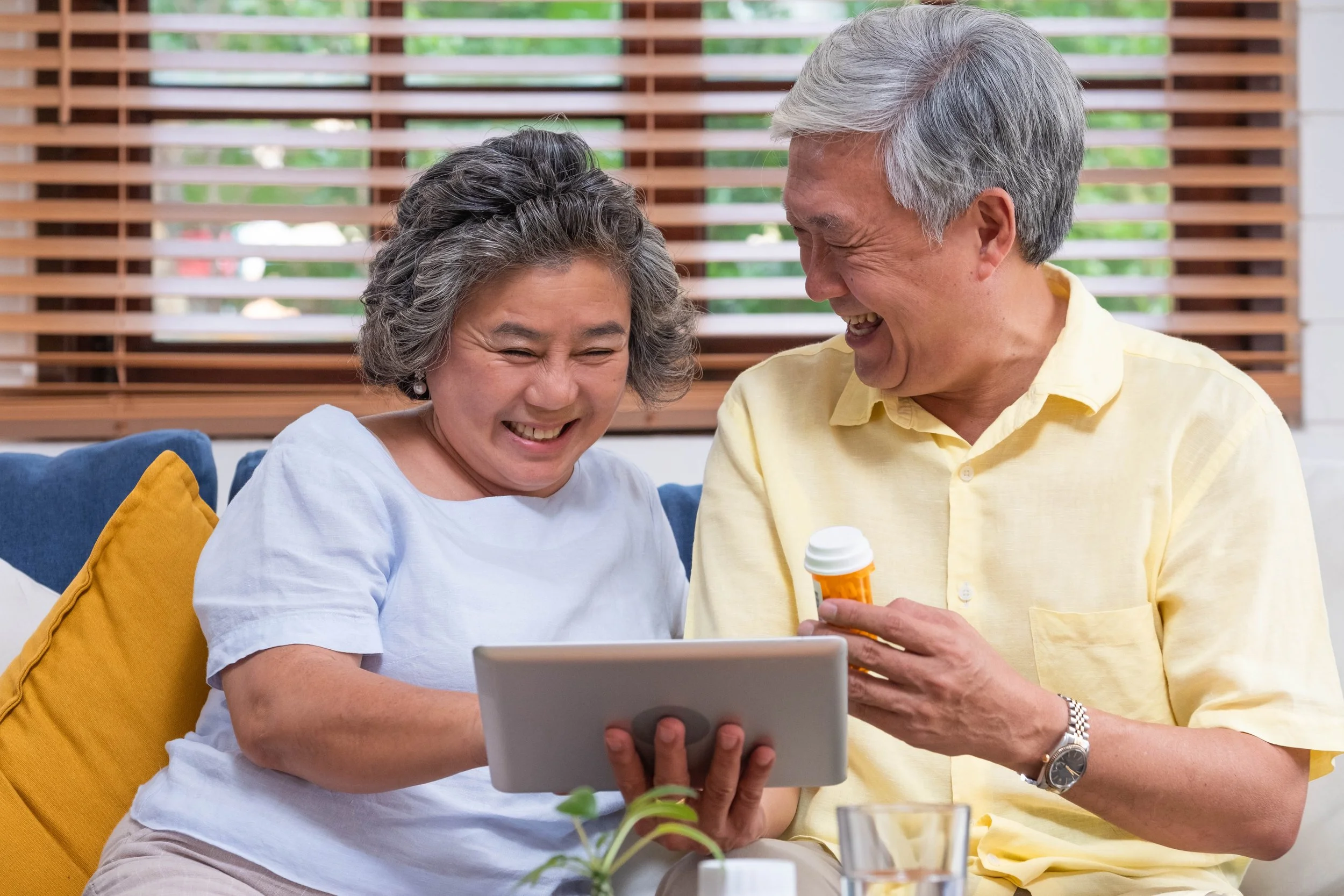 A smiling elderly woman and man sitting on a couch, looking at a tablet, with the man holding medication bottle, in a cozy home setting.
