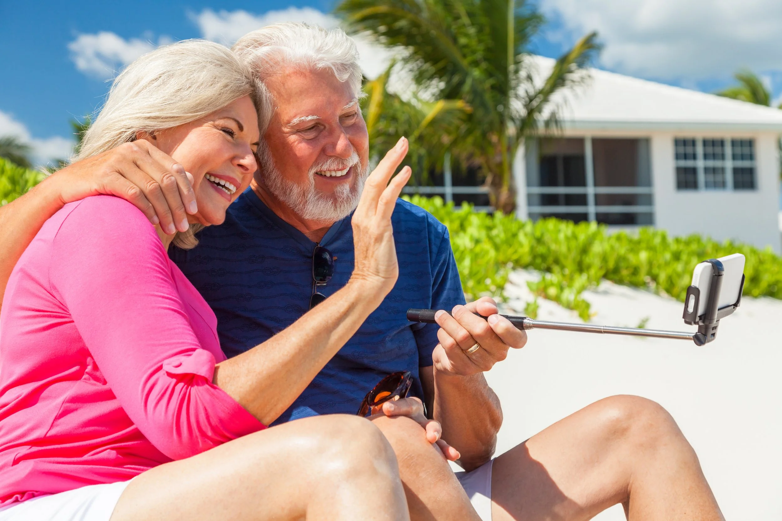 An elderly couple sitting outdoors on a sunny day, taking a selfie with a smartphone mounted on a selfie stick. The woman is wearing a pink shirt and the man is wearing a dark blue shirt with sunglasses hanging from his shirt. They are smiling and enjoying a moment together near a white house surrounded by greenery and palm trees.