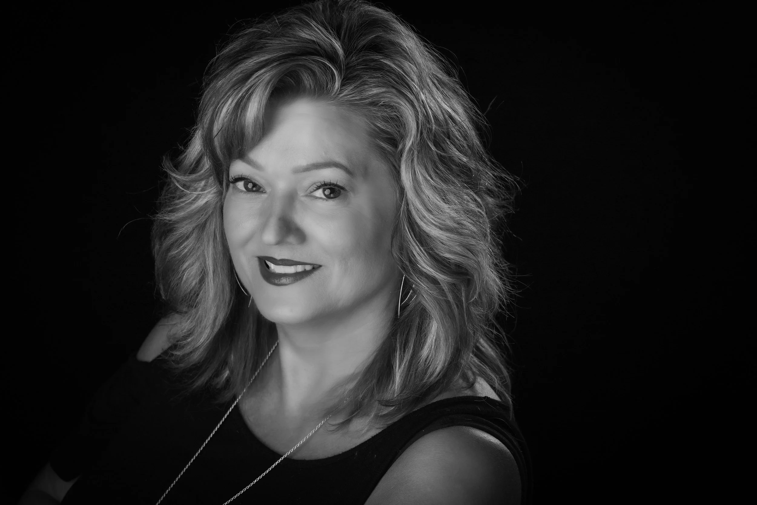Black and white portrait of a smiling woman with wavy hair wearing a dark top and necklace against a dark background.