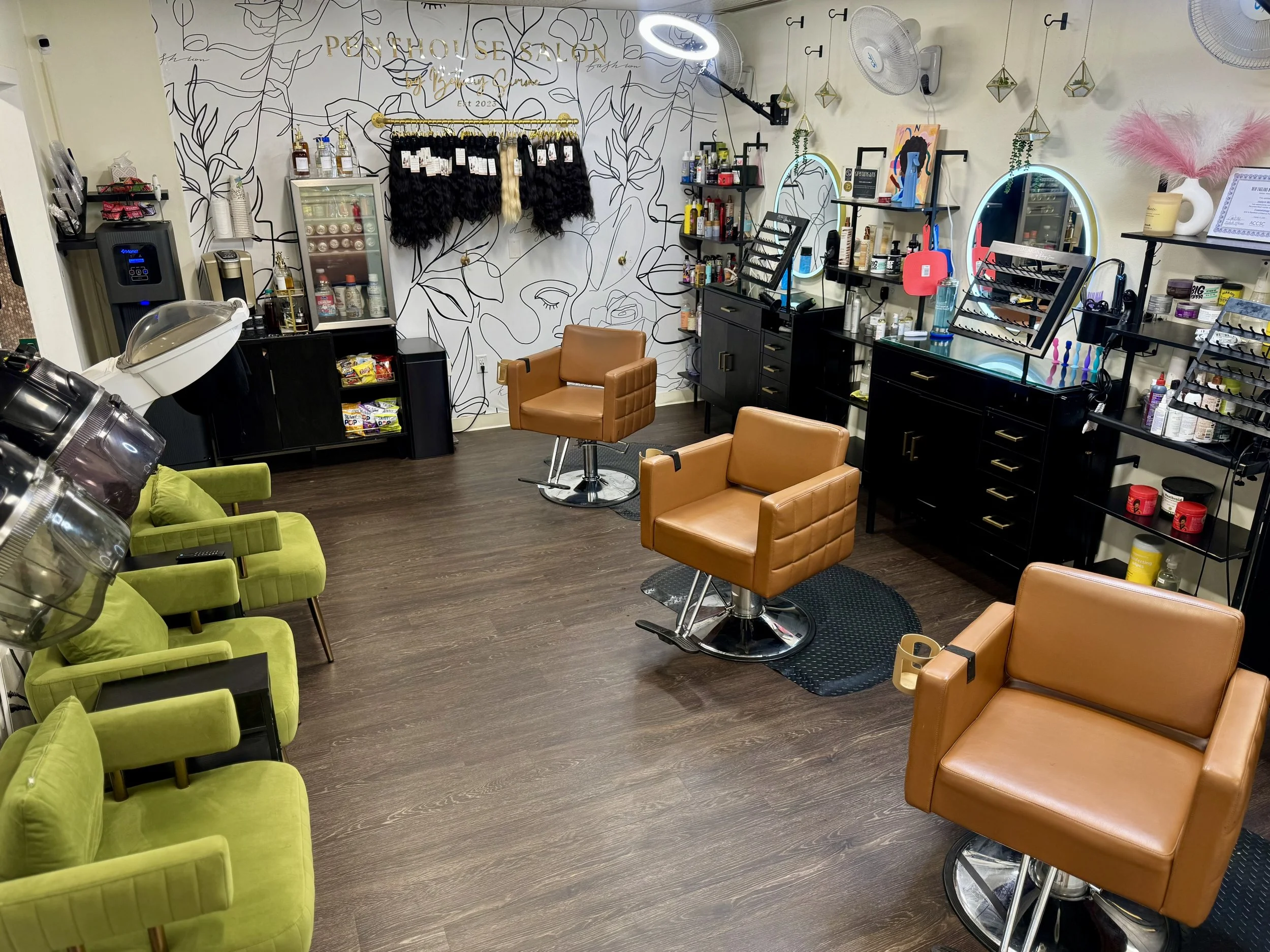 Interior of a hair salon with three brown chairs and three green chairs. The back wall features shelves with hair products, a mirror with lights, and business decor. The floor is dark wood, and there are hair styling tools and supplies visible on the shelves.