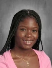 Young girl with braided hair wearing a pink top and a gold necklace with a cross pendant, smiling.