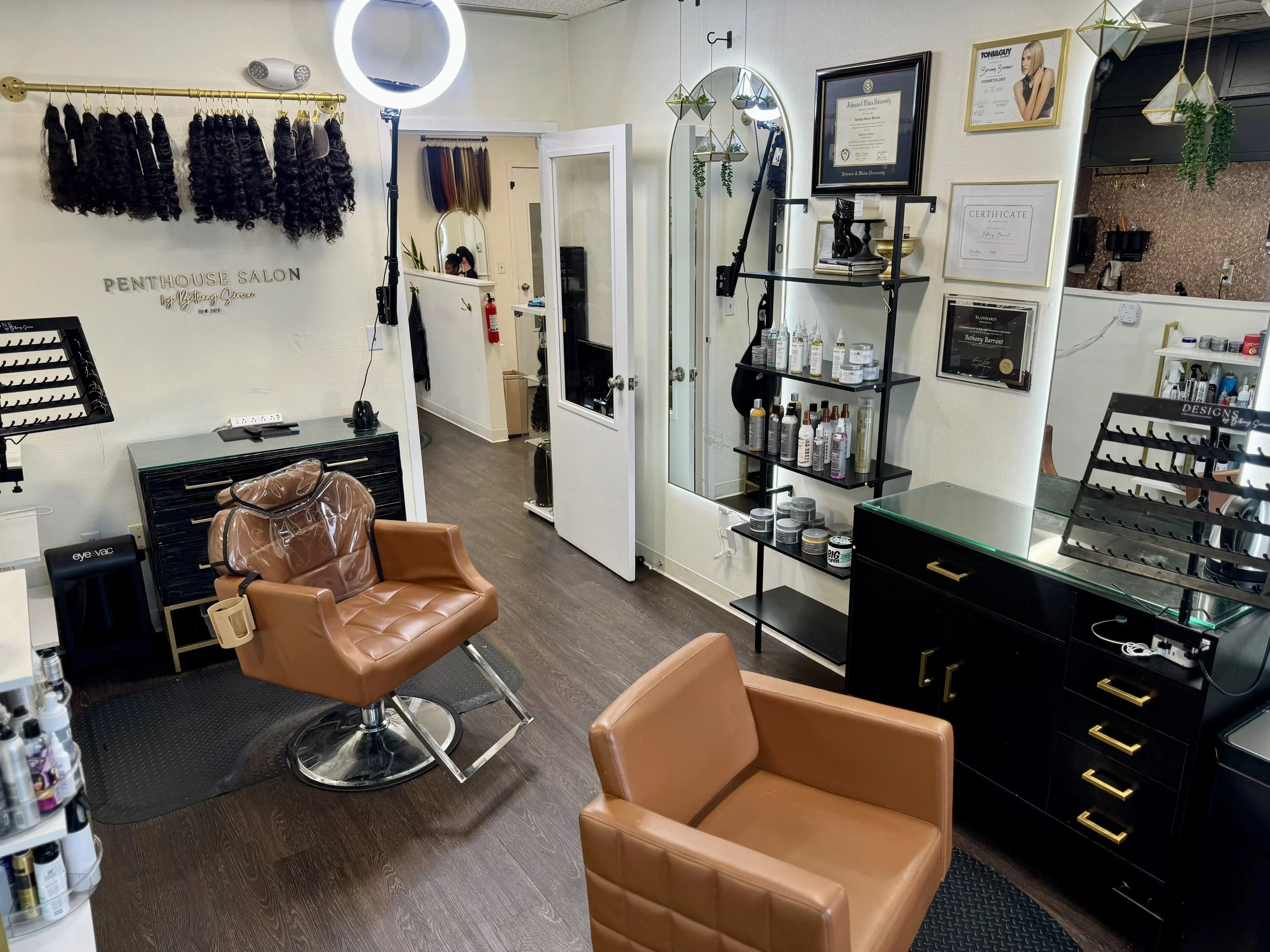 Interior of a hair salon with a styling chair, shelves with hair products, framed certificates and awards, and hair extensions hanging on the wall.