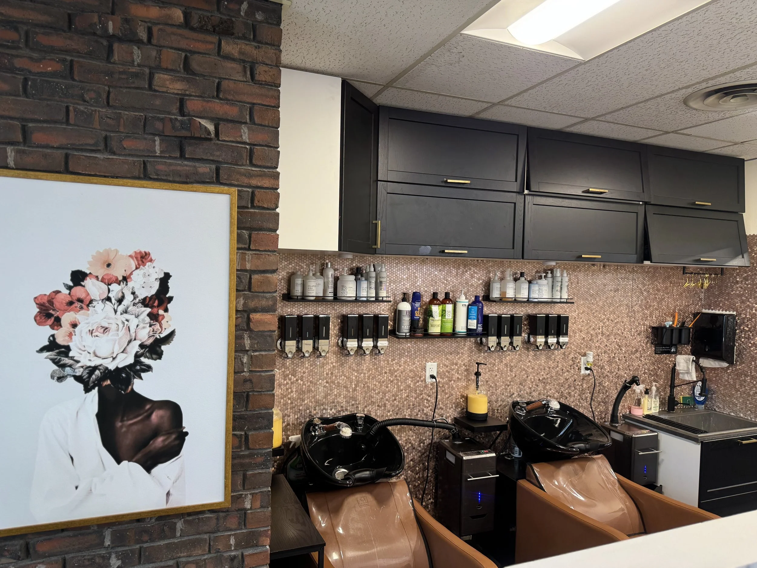 Interior of a hair salon featuring two shampoo stations with black sinks, brown chairs, wall-mounted shelves with various hair care products, black cabinetry, and a framed art piece depicting a person with a floral head covering, set against a brick wall.