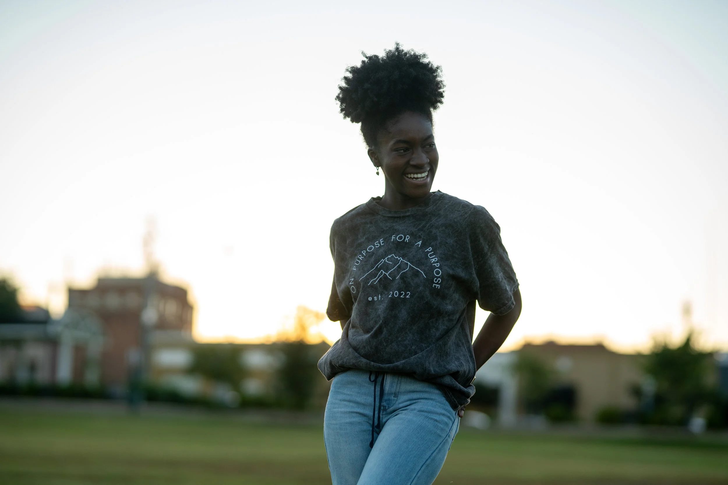 A smiling Black woman with curly hair standing outdoors during sunset, wearing a dark t-shirt with mountain and text design and light blue jeans.