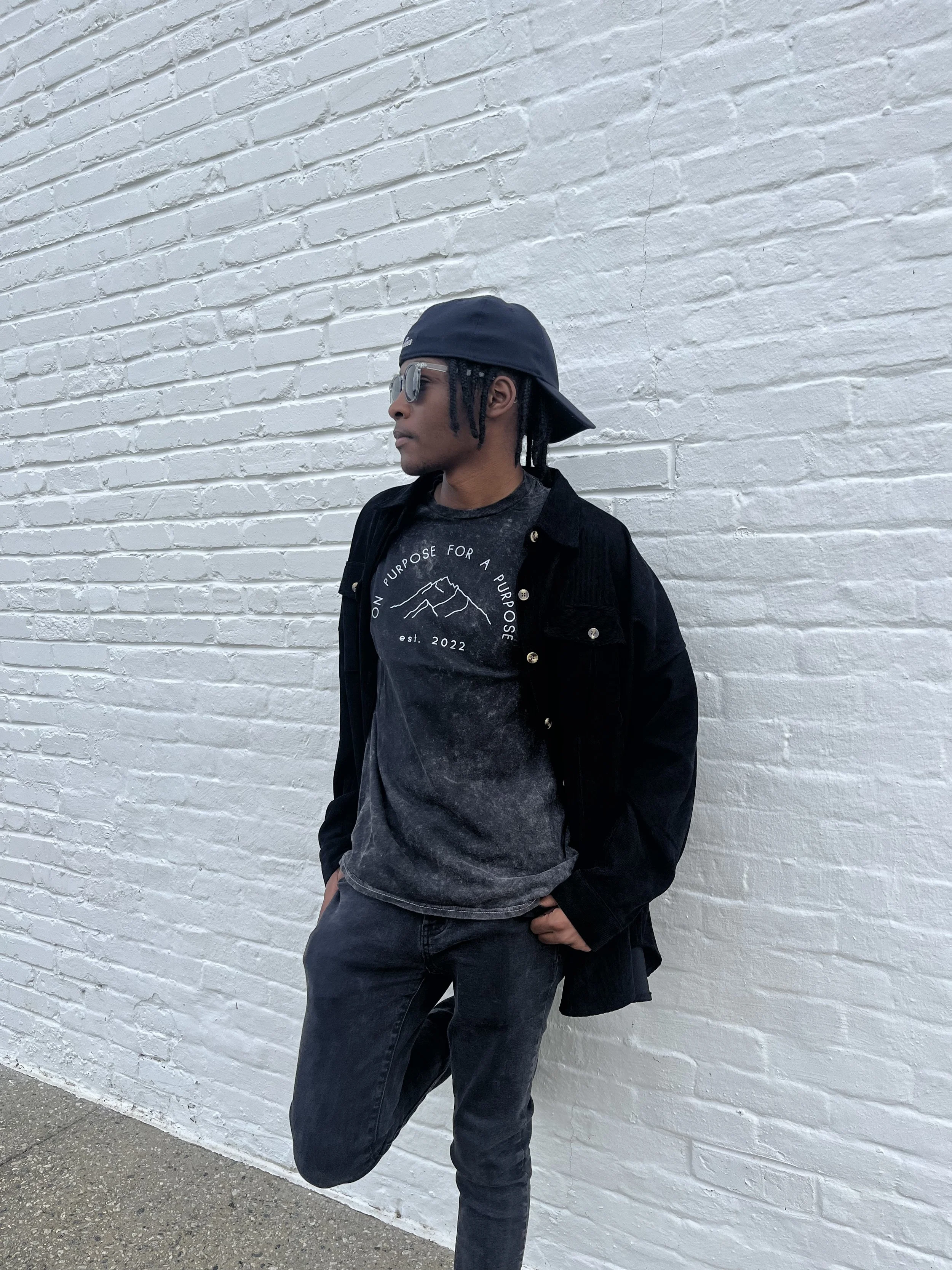 A young man wearing a backwards black cap, sunglasses, a black jacket, and a black graphic t-shirt, leaning against a white brick wall with one foot up on the wall.