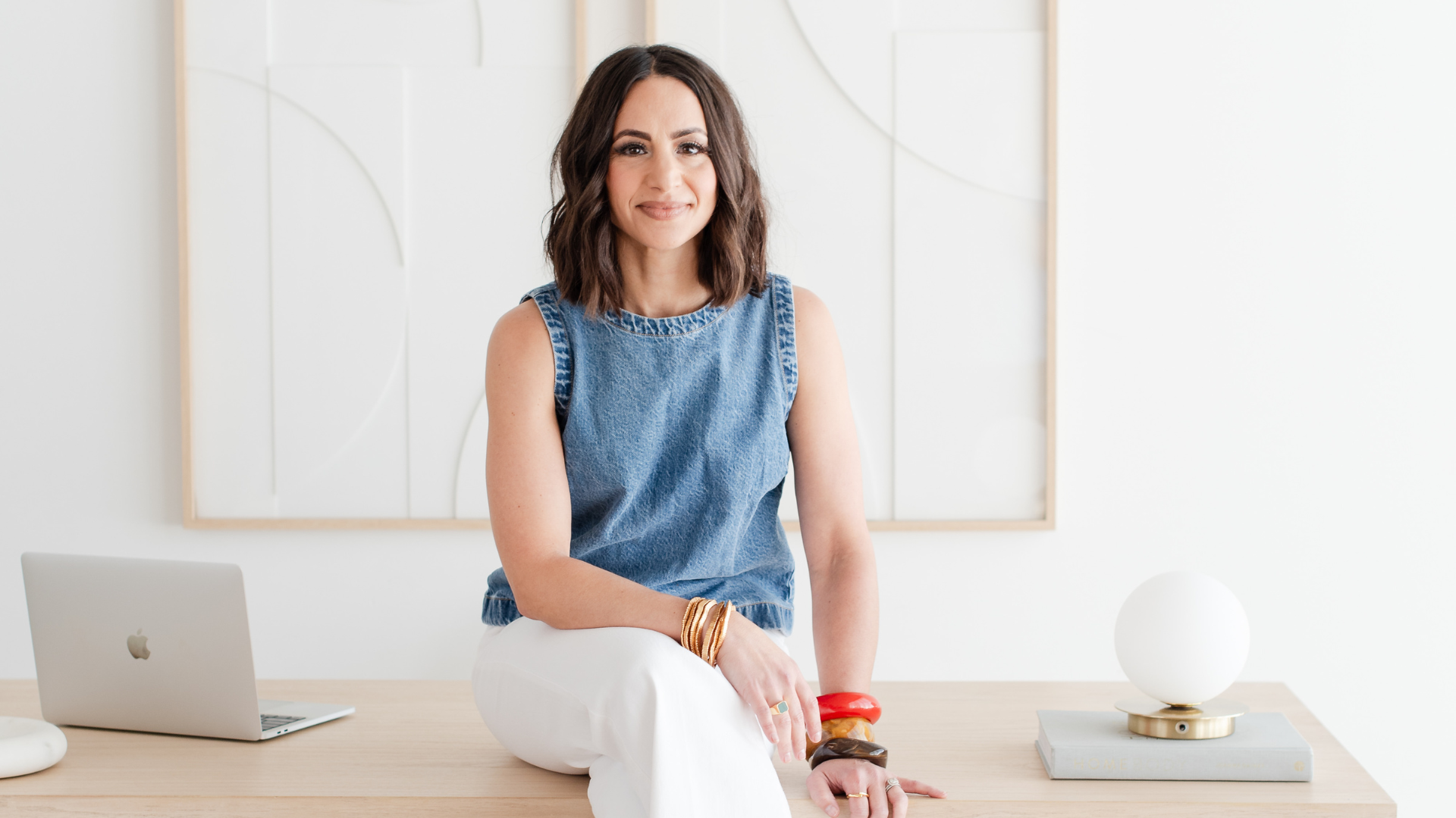 A woman with medium-length brown hair, wearing a sleeveless denim top and white pants, sitting on a wooden desk in a modern, minimalistic room. There is an Apple laptop to her left and a white spherical lamp on the right. She is smiling softly with her hands resting on her knee, wearing multiple gold bracelets and a ring.