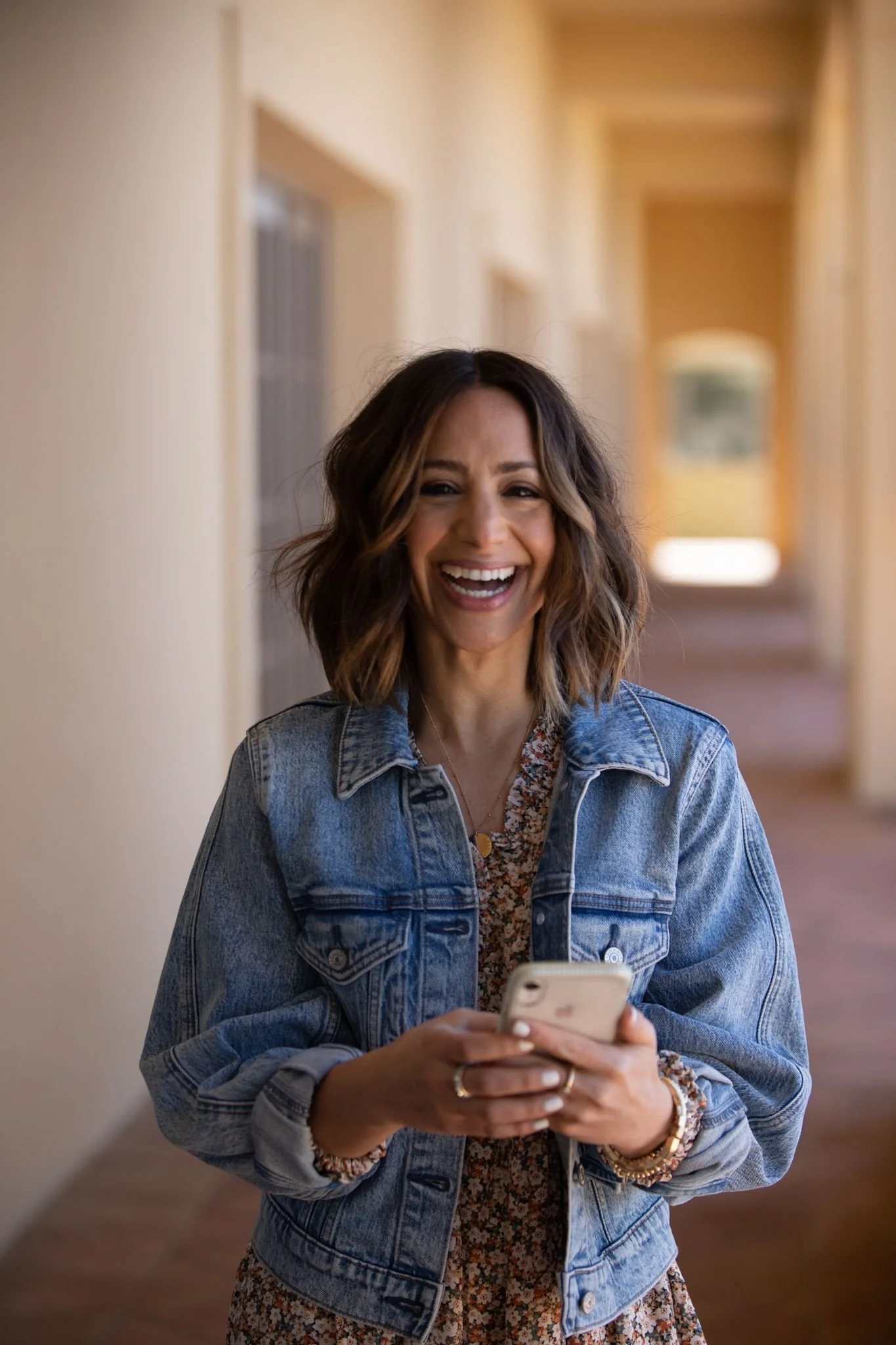 A woman with shoulder-length wavy brown hair, wearing a denim jacket over a floral dress, smiling and looking at her phone while standing in a hallway.