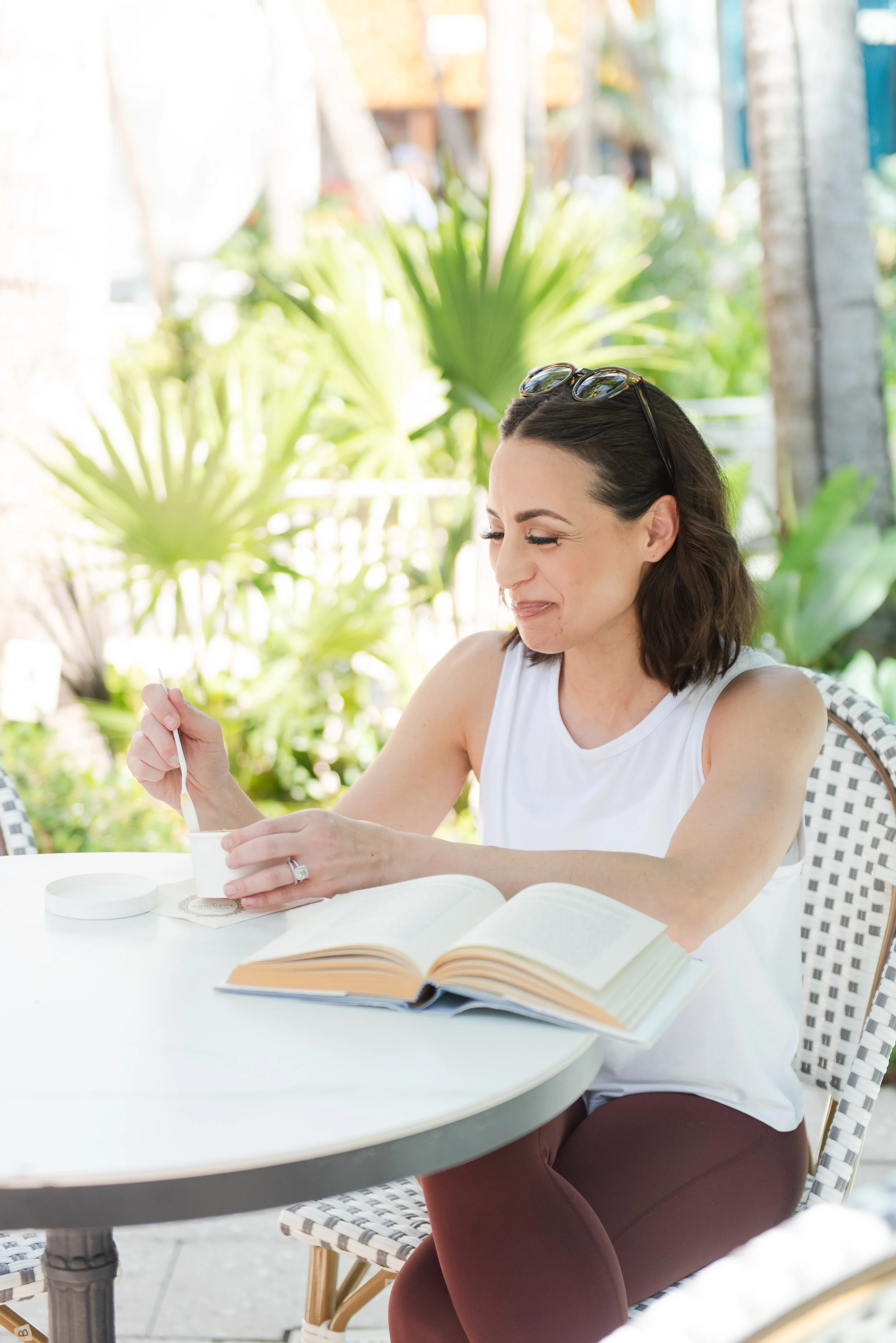 A woman with dark hair, glasses on her head, wearing a white tank top, is sitting at an outdoor table, smiling and eating from a bowl with a spoon. There is an open book on the table, and she has a diamond ring on her left hand. The background has lush green plants and trees.