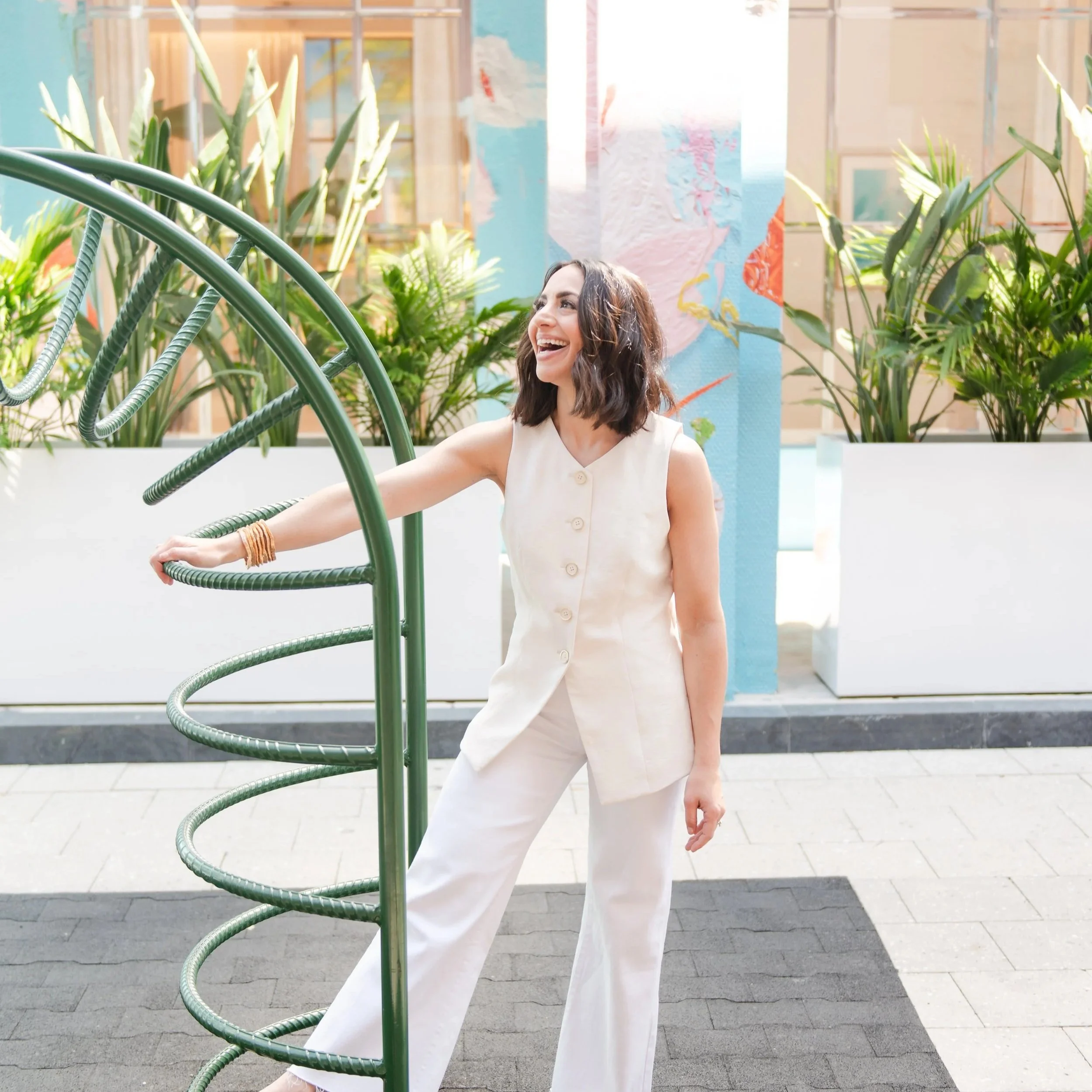 A woman with dark hair, wearing a sleeveless cream-colored dress, is smiling and sitting near a spiral-shaped outdoor staircase, surrounded by green tropical plants, with a colorful artwork on a blue wall in the background.