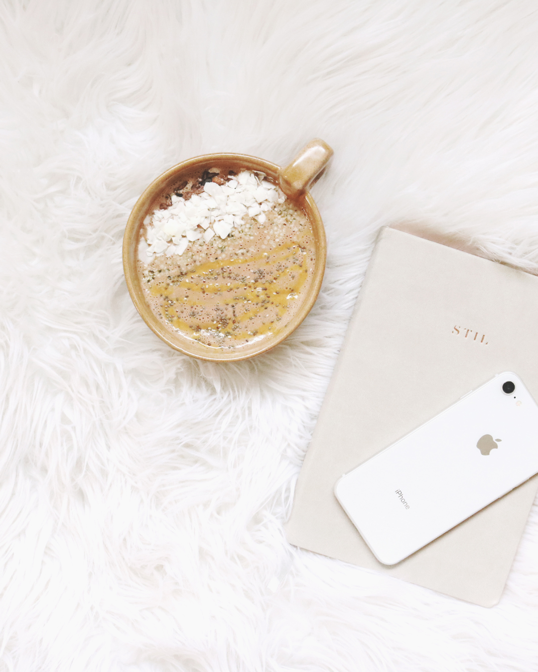 A beige mug filled with a beverage topped with white chocolate shavings, chia seeds, and caramel drizzle, placed on a white fluffy surface next to a white iPhone and a beige notebook.