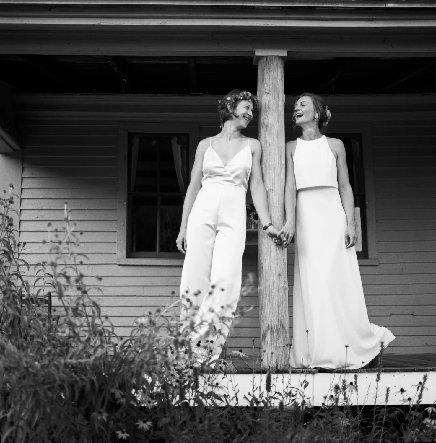 A black and white film photograph of two brides (one bride is wearing a pants suite) holding hands standing on a rustic wooden porch with wildflowers in the foreground.