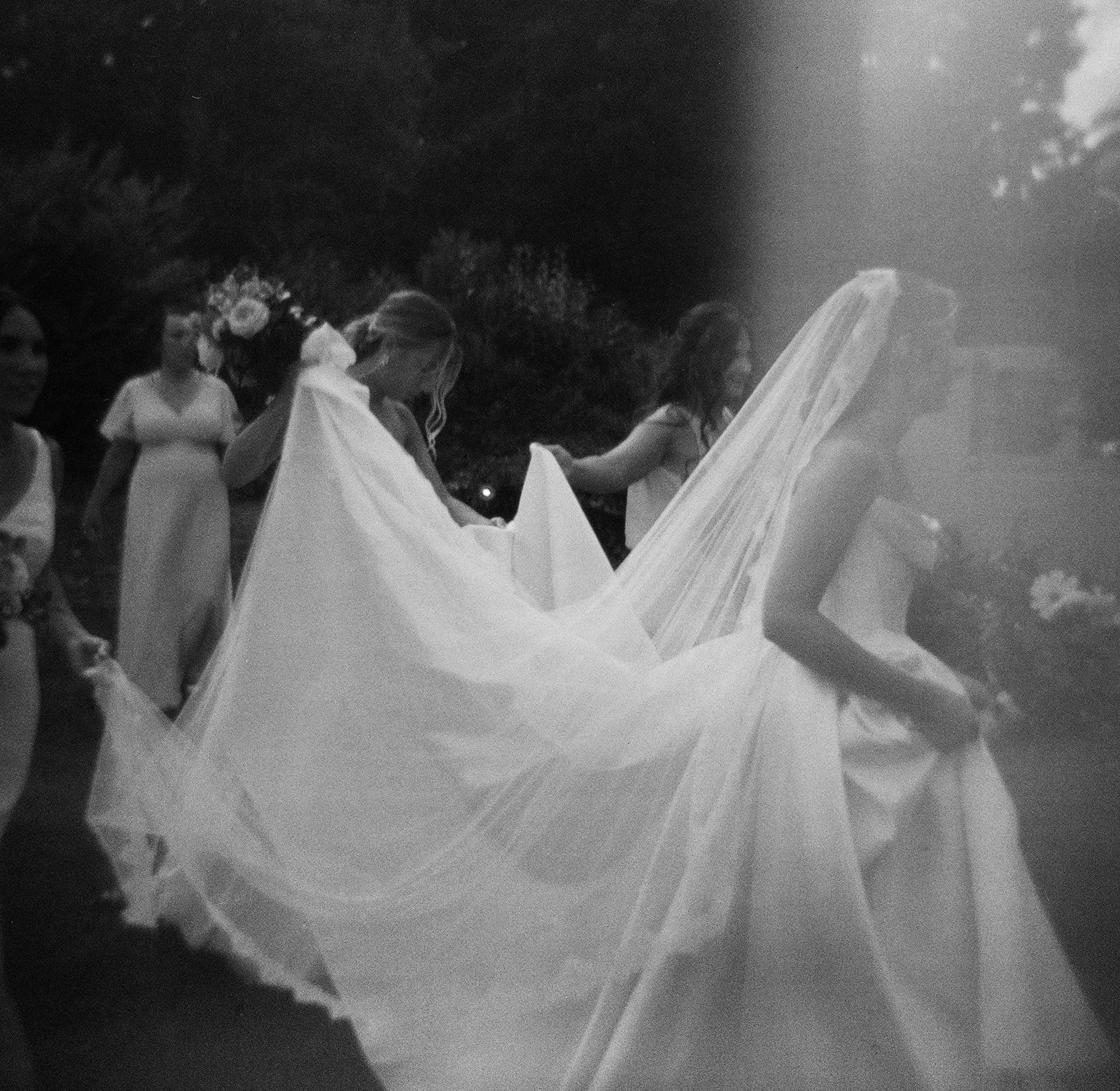 A black and white film photograph of a bride with her bridesmaids helping her with her dress and viel. The photo has a romatic, soft, vintage look. The wedding party is at Misselwood in Beverly, MA., photo by Christina Richards in Boston.