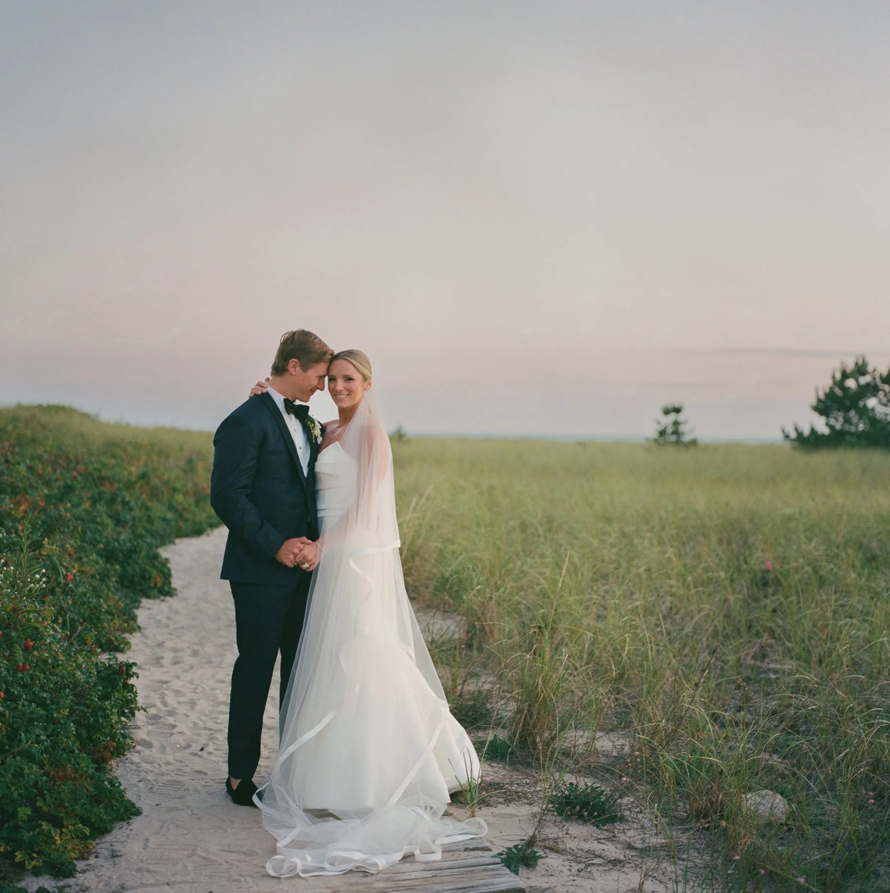Wychmere Beach Club wedding; the couple is smiling at the camera with a hazy background while embracing on the dunes. Color film photo by Christina Richards