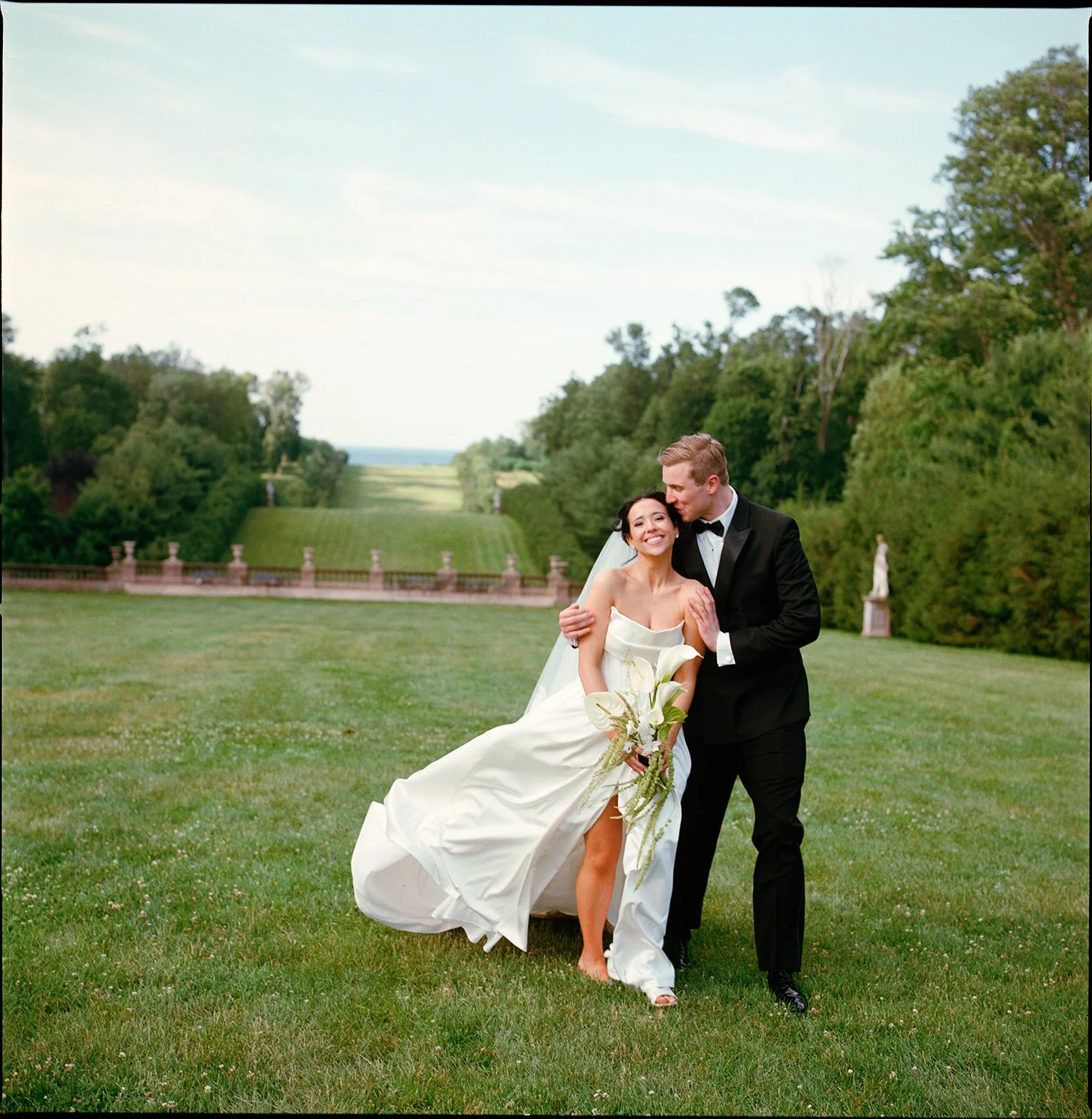 A wedding photo of a couple at the Crane Estate with the rolling hills that overlooks the ocean in the background. The couple looks happy, the groom hugs the bride and kisses her as she smiles. The photo has a dreamy quality and is shot on color film