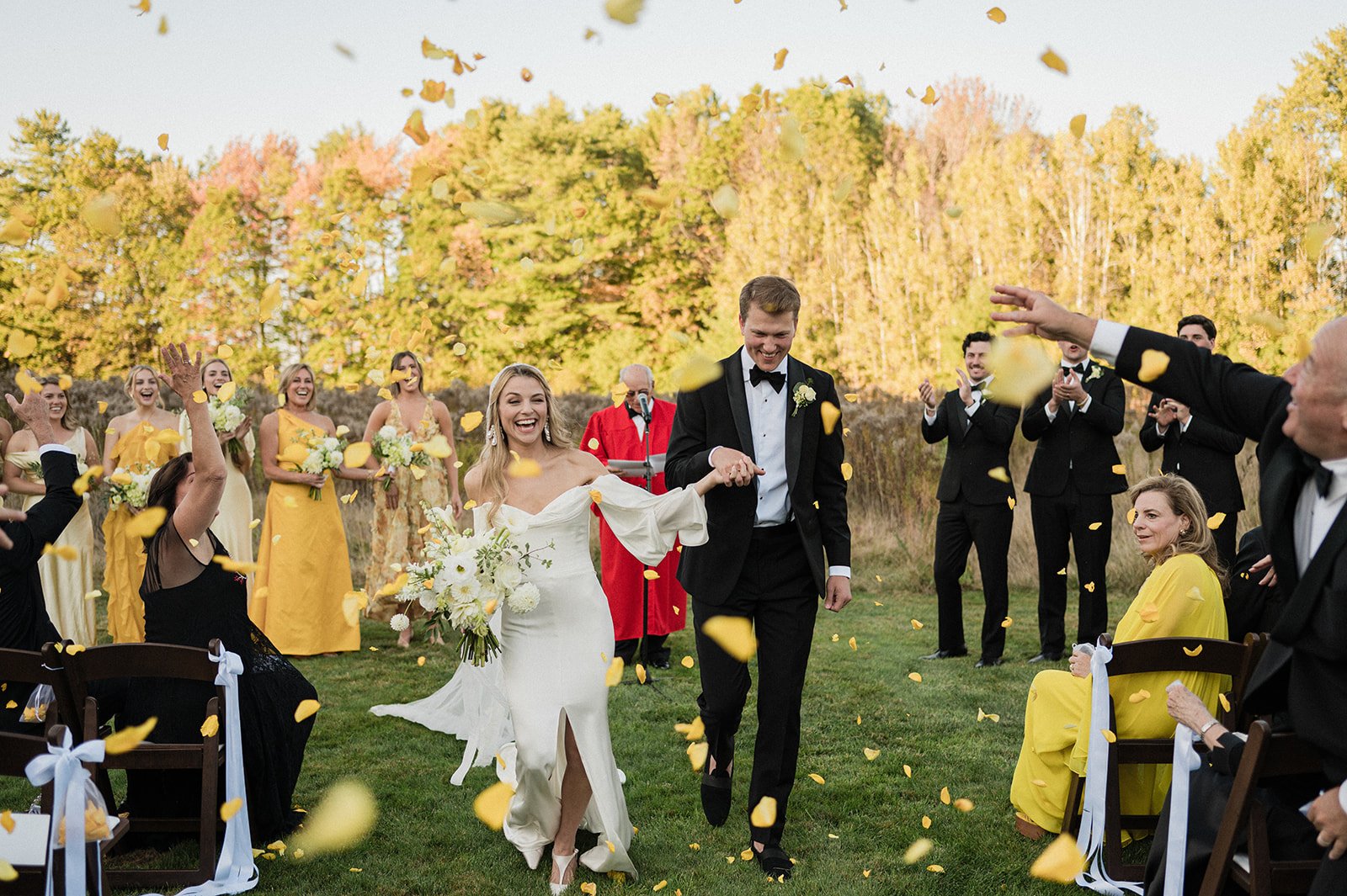 A color documentary wedding photograph at Flanagan Farm in Maine. A wedding couple is smiling as they exit thier wedding ceremony as guests toss yellow flower petals. The photo is by Boston based wedding photographer, Christina Richards
