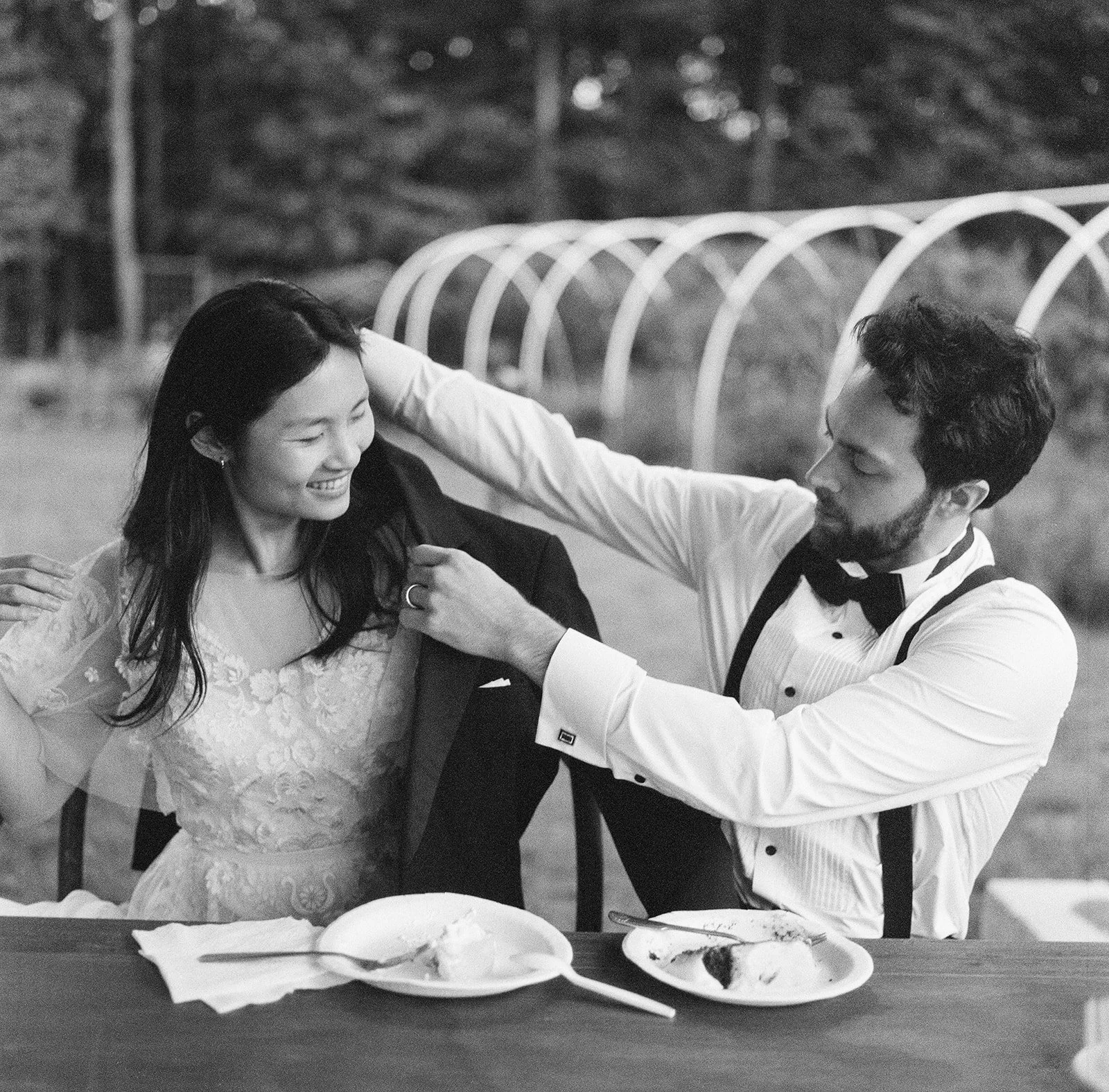 A black and white film photograph of a groom putting his jacket on the bride during a backyard wedding reception.