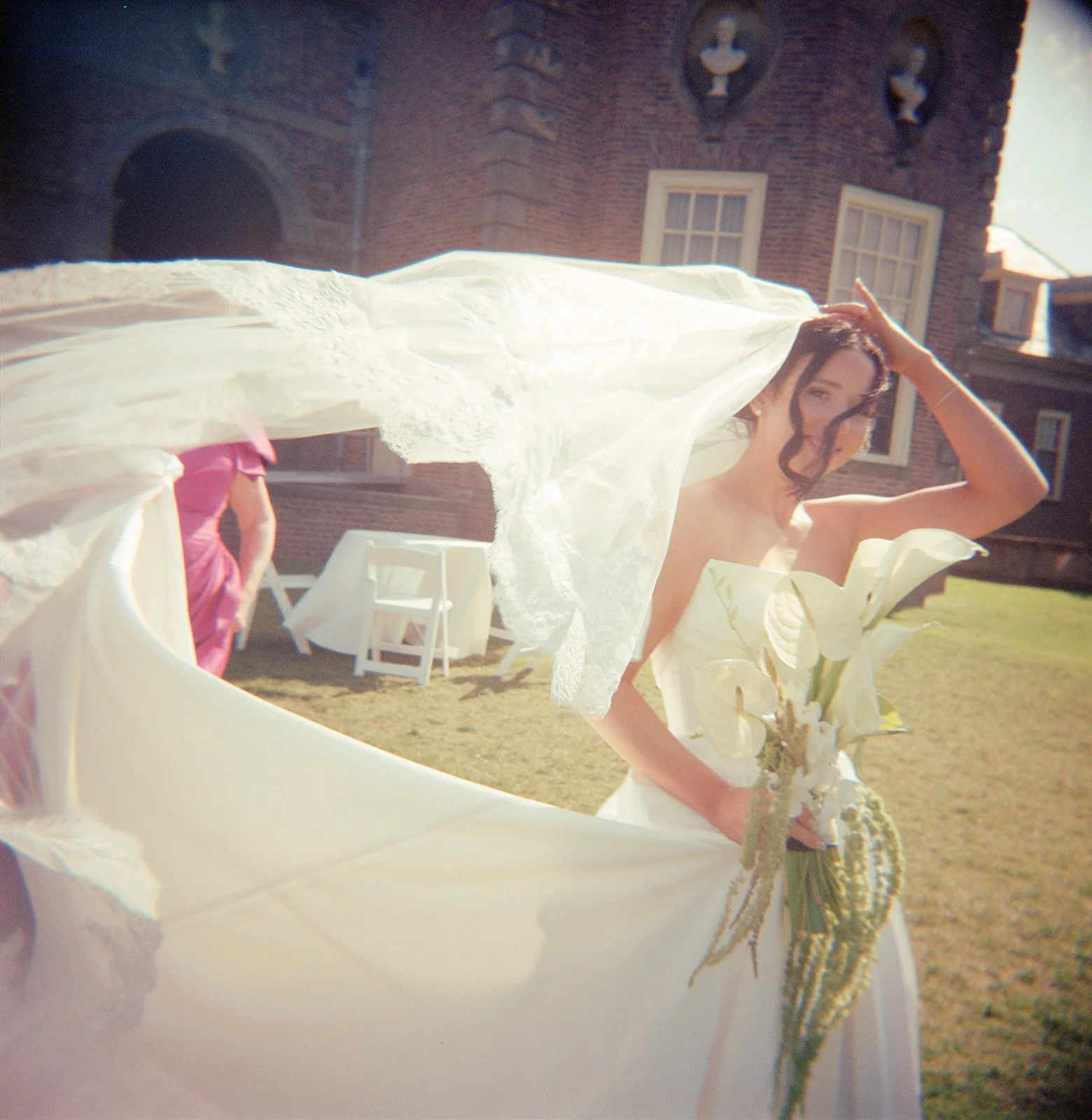 Color film photo of a bride on a windy day at the Crane Estate. The bride holds the top of her veil and as it flutters in the wind. The bride is holding white lily flowers in her bouquet. The photo has a dreamy, soft, artistic, look. By Christina Ric