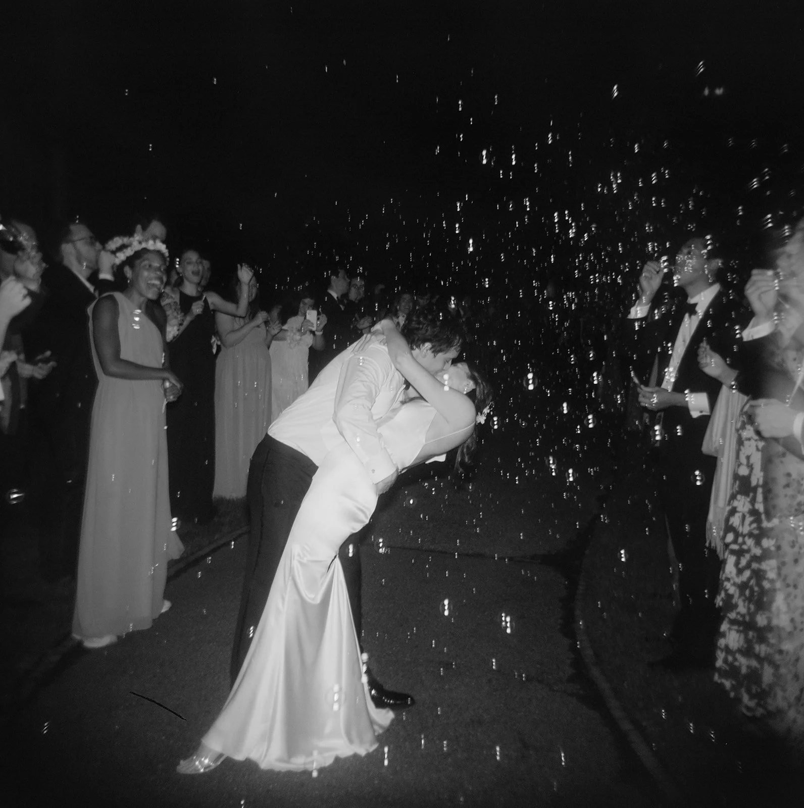 A black and white film wedding photograph of a wedding couple doing a dip kiss during a bubble exit at the end of the wedding reception at Harvard. The photo has a vintage, fun,  and artistic feel. By bosed based photographer Christina Richards