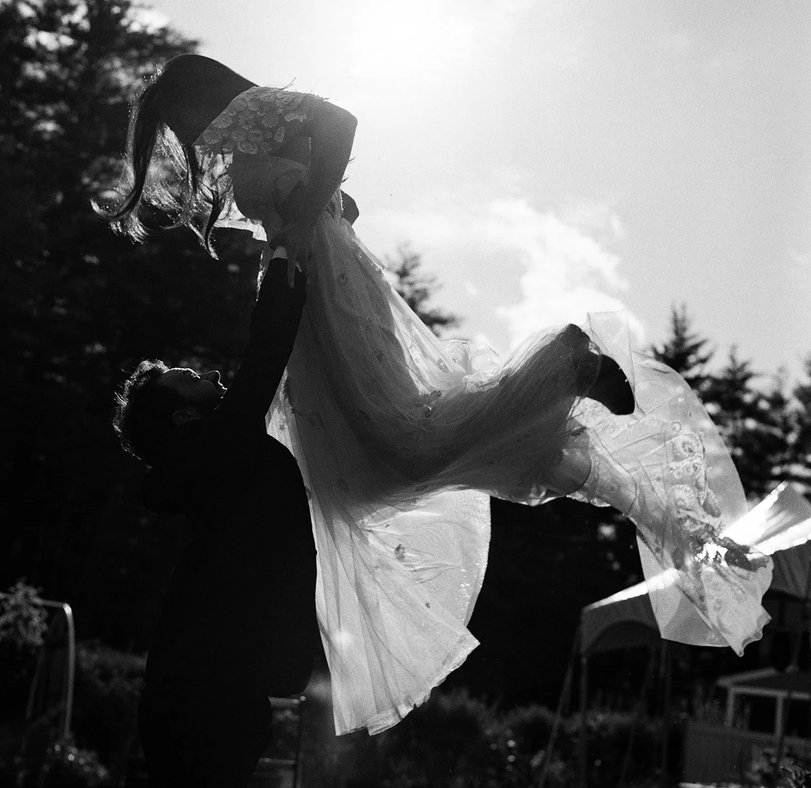 A black and white artistic film photograph of a groom lifting the bride in the air after thier backyard wedding ceremony.