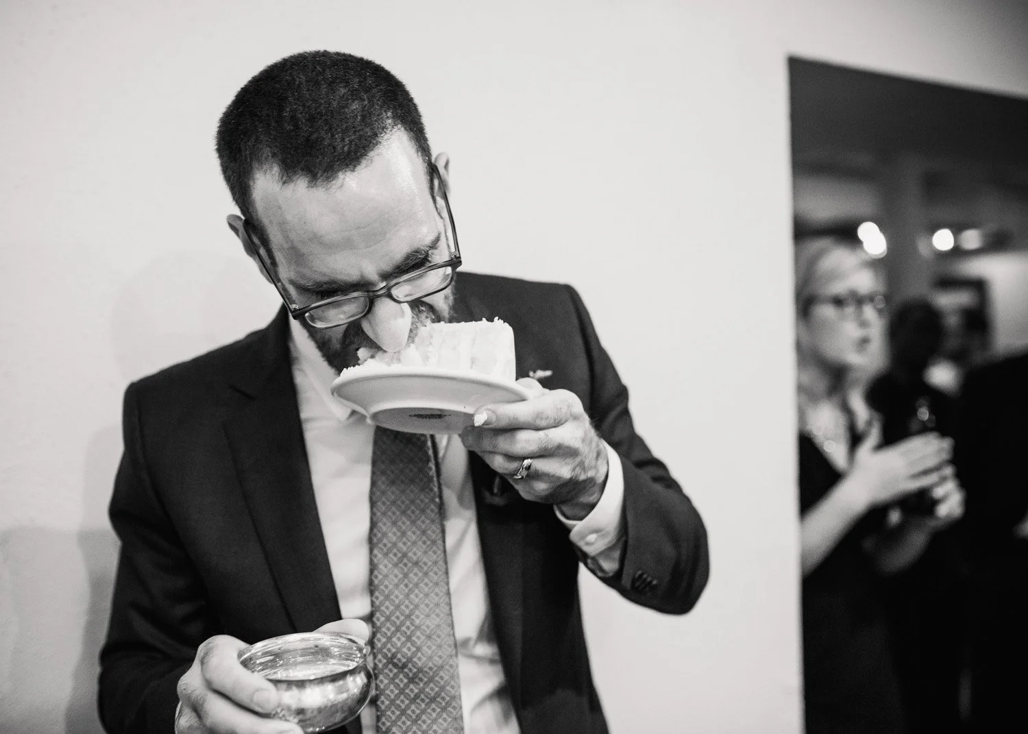 A black and white documentary wedding photograph of a groom eating his wedding take without silverware. The photograph is candid and fun.