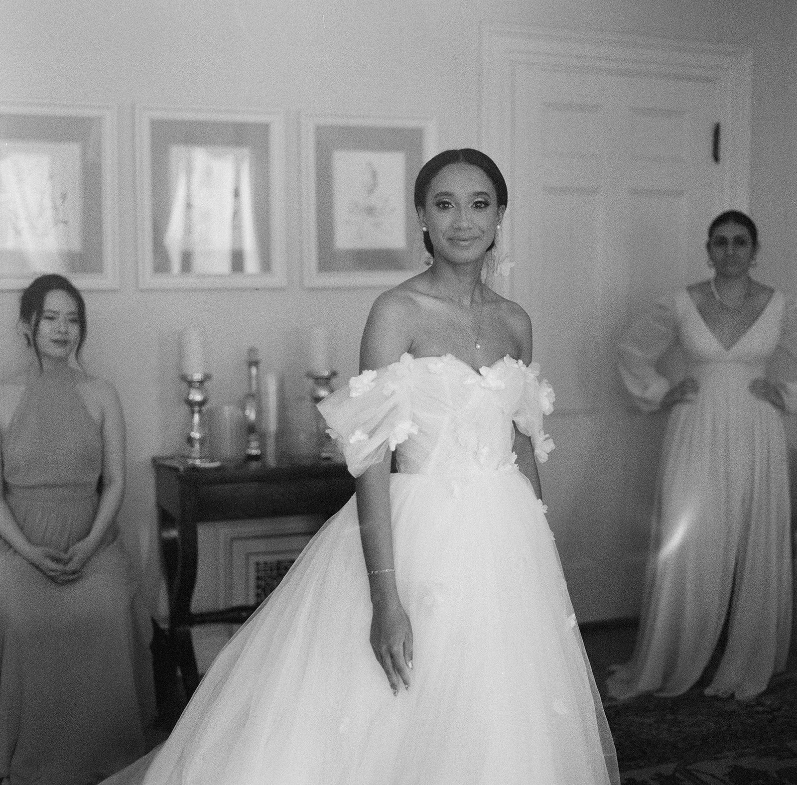 A black and white film photo of a bride centered in the frame with her bridesmaids on the edge of the frame. The bride is waiting in the bridal suite of Glen Magna Farm. Photo by Boston based Christina Richards.