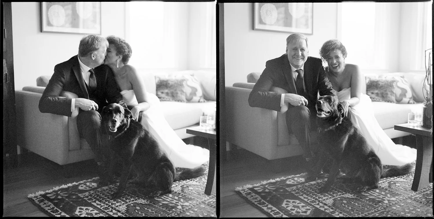 Black and white film wedding photo of a couple sitting on their couch at home with their dog. They are kissing, then laughing. The photo has an artistic vibe, photographed by Boston based Christina Richards.  