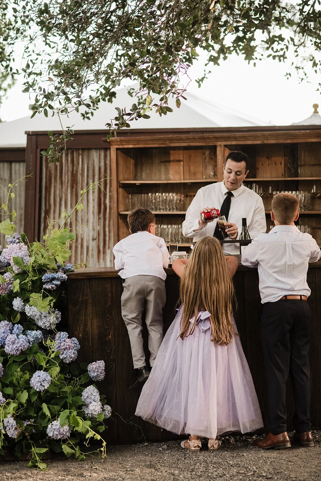 The flower girl and ring bearers gather at the outdoor bar at Arista Winery decorated with hydrangeas. A candid humorous wedding photo.