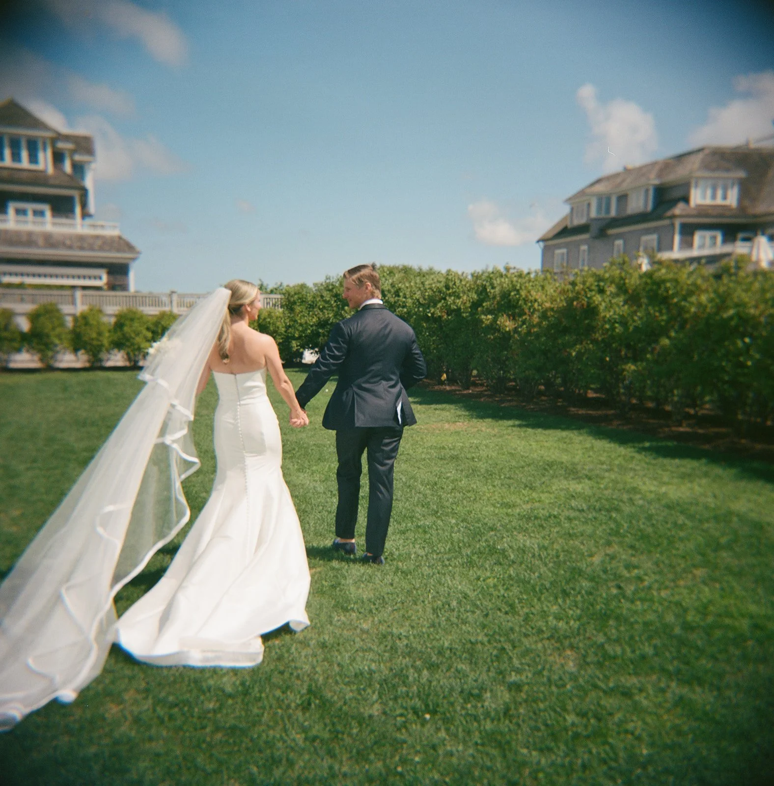 Bride and groom on the green lawn of Wychmere in Cape Cod walking away from the camera. Shot on film, the image has a soft vintage look.