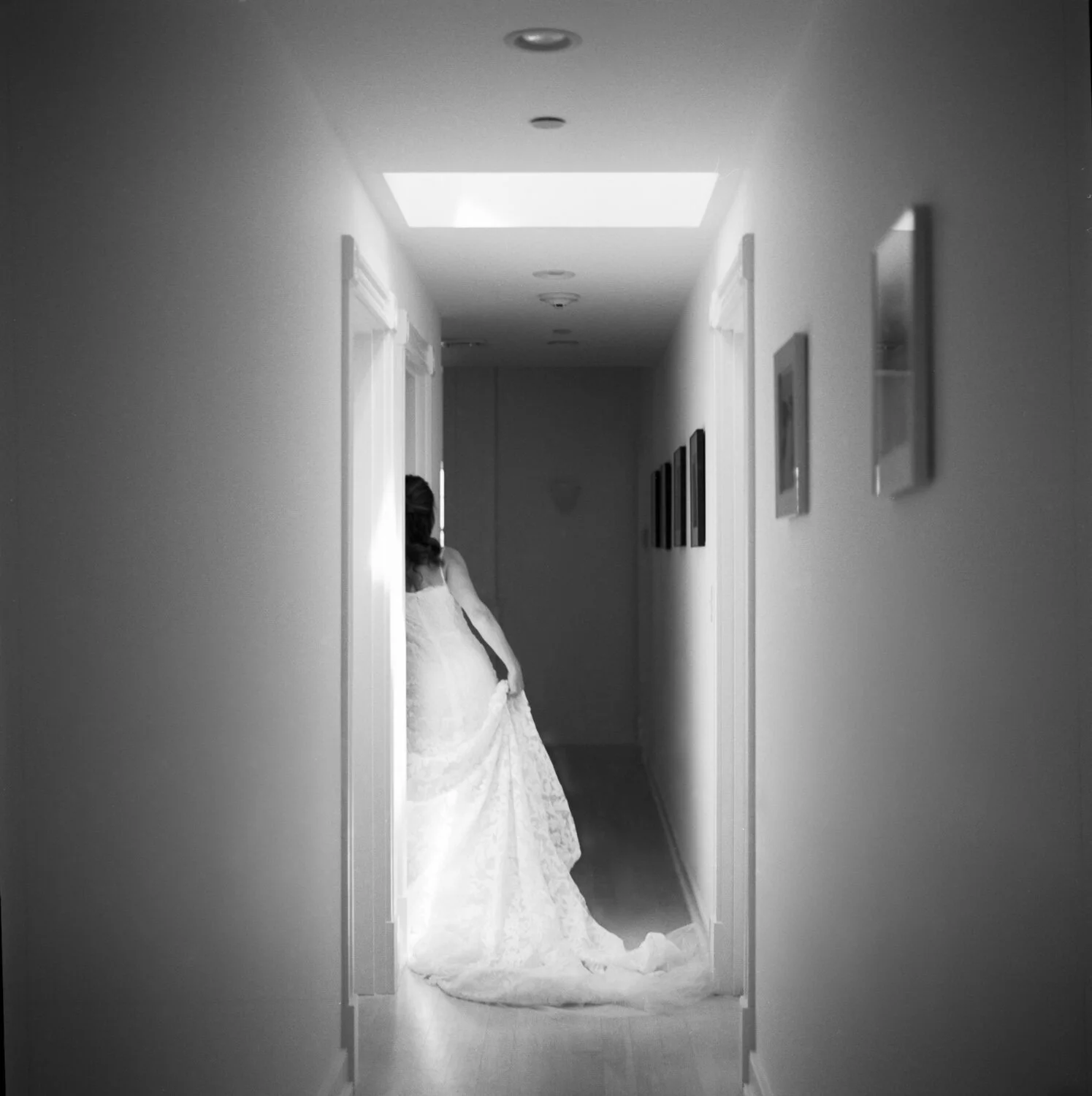 A black and white film photo of a bride getting ready at a hotel in Nantucket. A skylight gives the photo a glow from above. The bride is gently pulling on her dress in an elegant way. By Christina Richards of Boston.