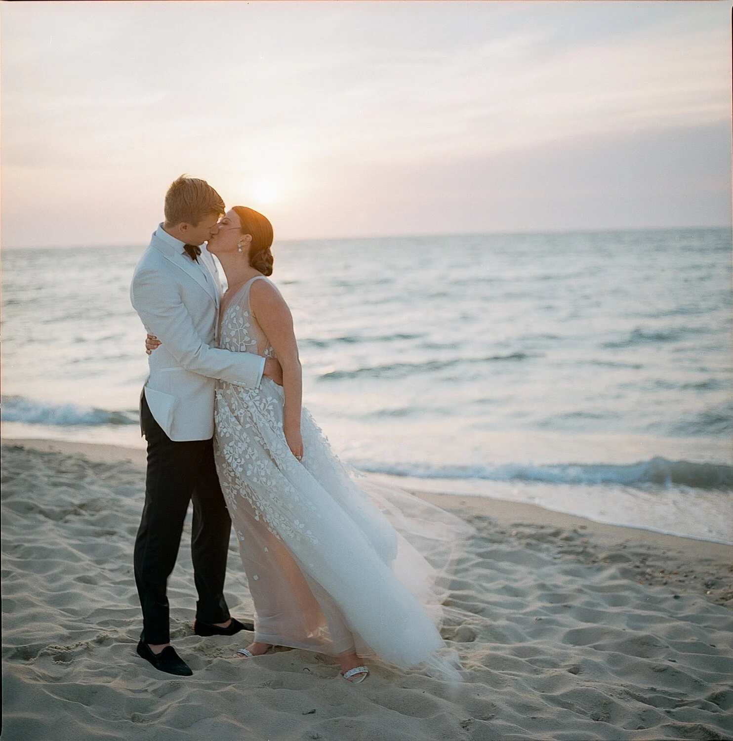 A color film photograph of a wedding couple kissing on the beach at sunset in Nantucket. The colors are soft and inviting.