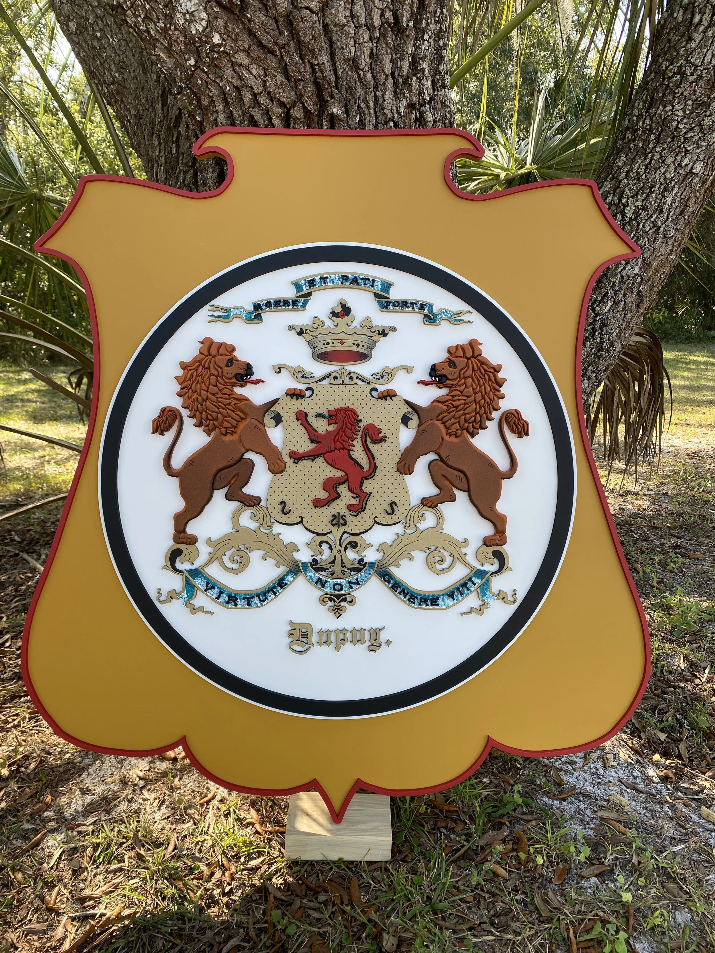 A gold shield-shaped sign with a coat of arms featuring two lions standing on their hind legs, holding a shield with a red lion, topped with a crown, and adorned with banners and decorative designs, positioned outdoors near trees and grass.