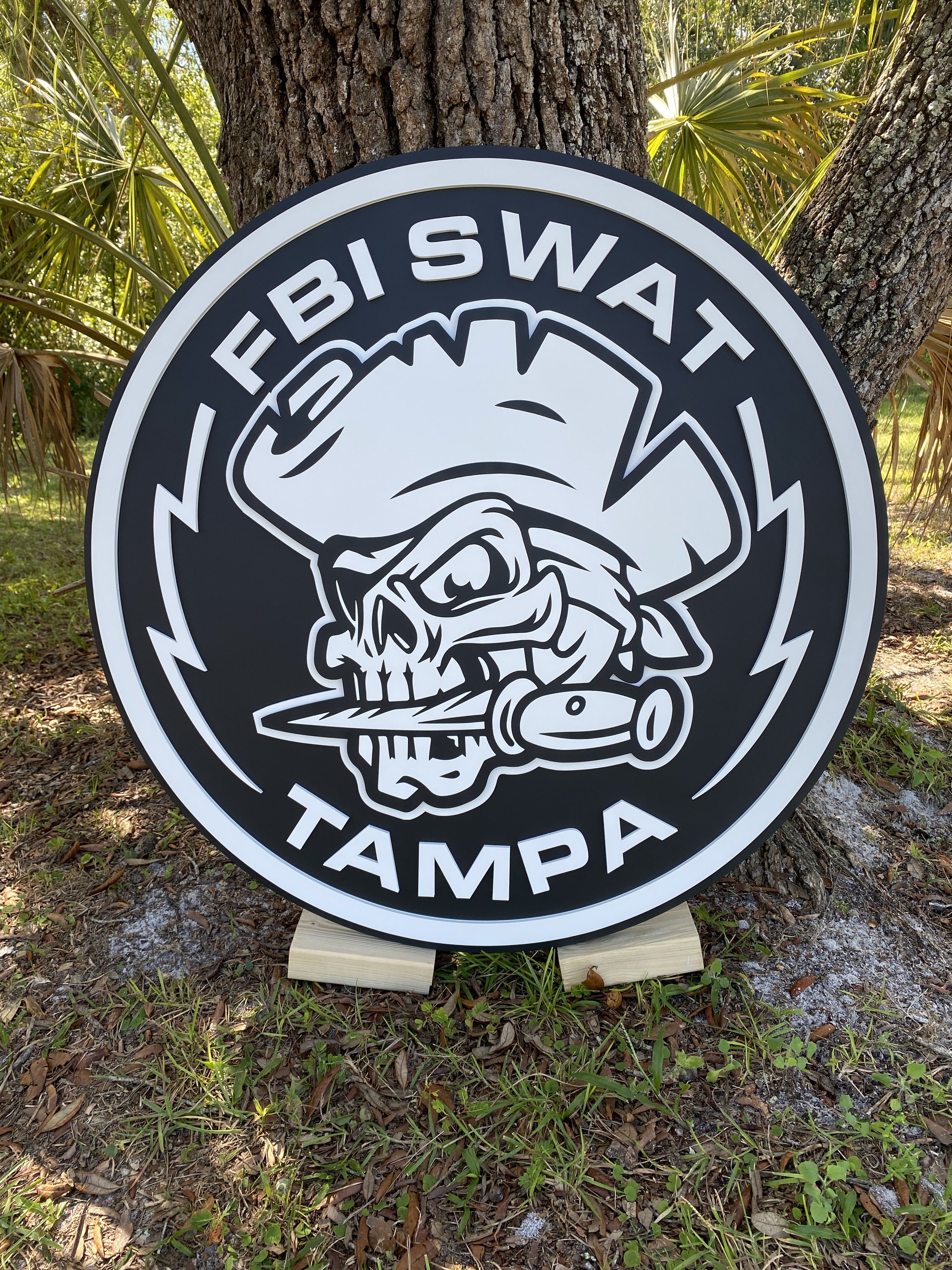 Signboard with skull wearing a pirate hat and eye patch, holding a dagger and a crossbow, with the text 'FBI SWAT TAMPA' around it, placed outdoors among trees and grass.