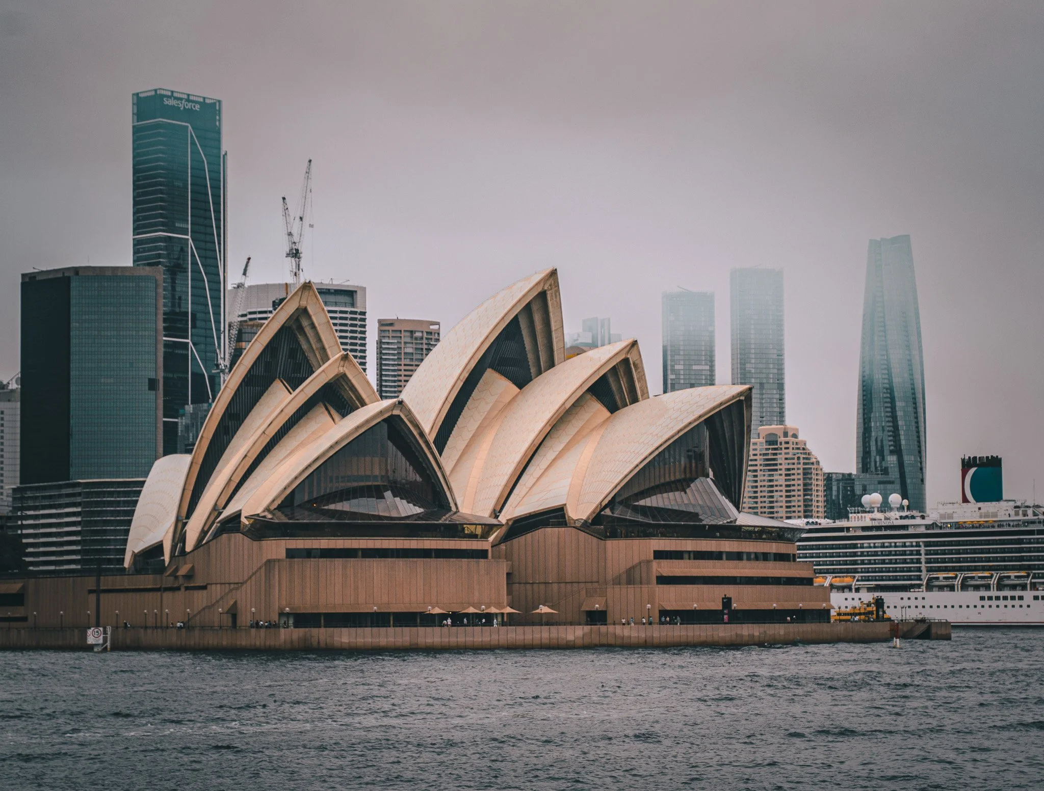 Sydney Opera House with modern skyscrapers in the background on a cloudy day, situated beside the water.